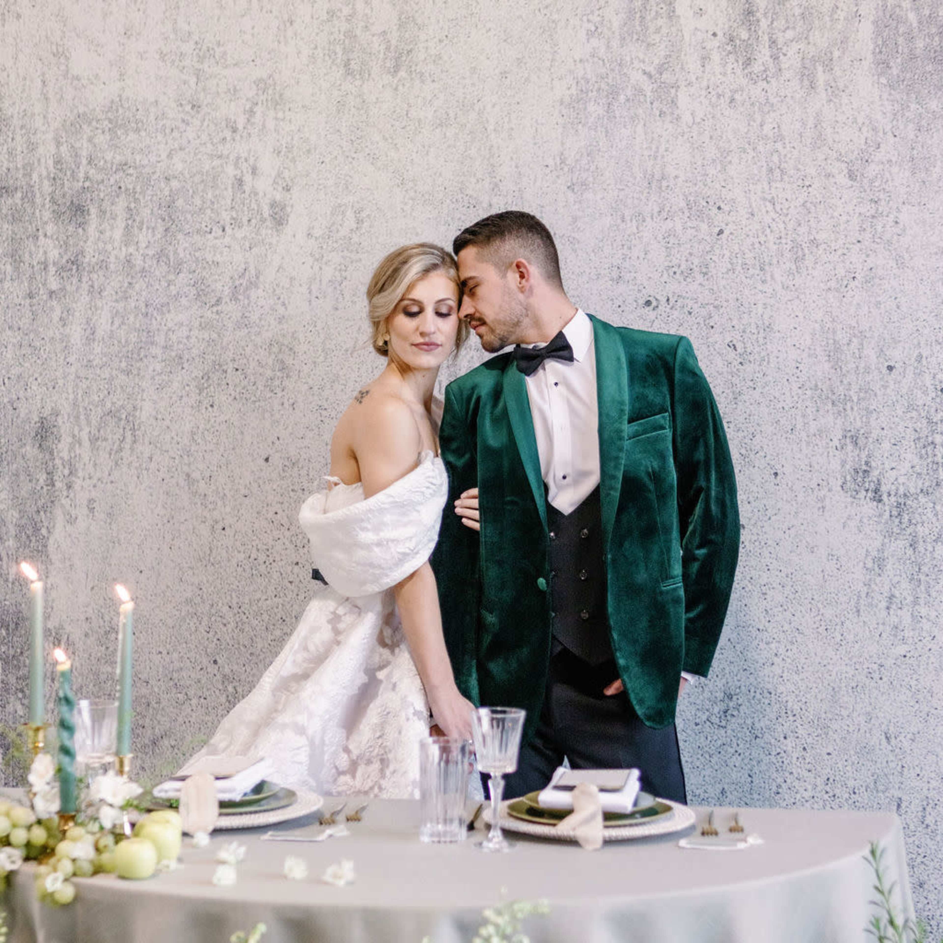 A couple stands together at an elegantly set table adorned with greenery and candles, with the woman in a wedding dress and the man in a green tuxedo.