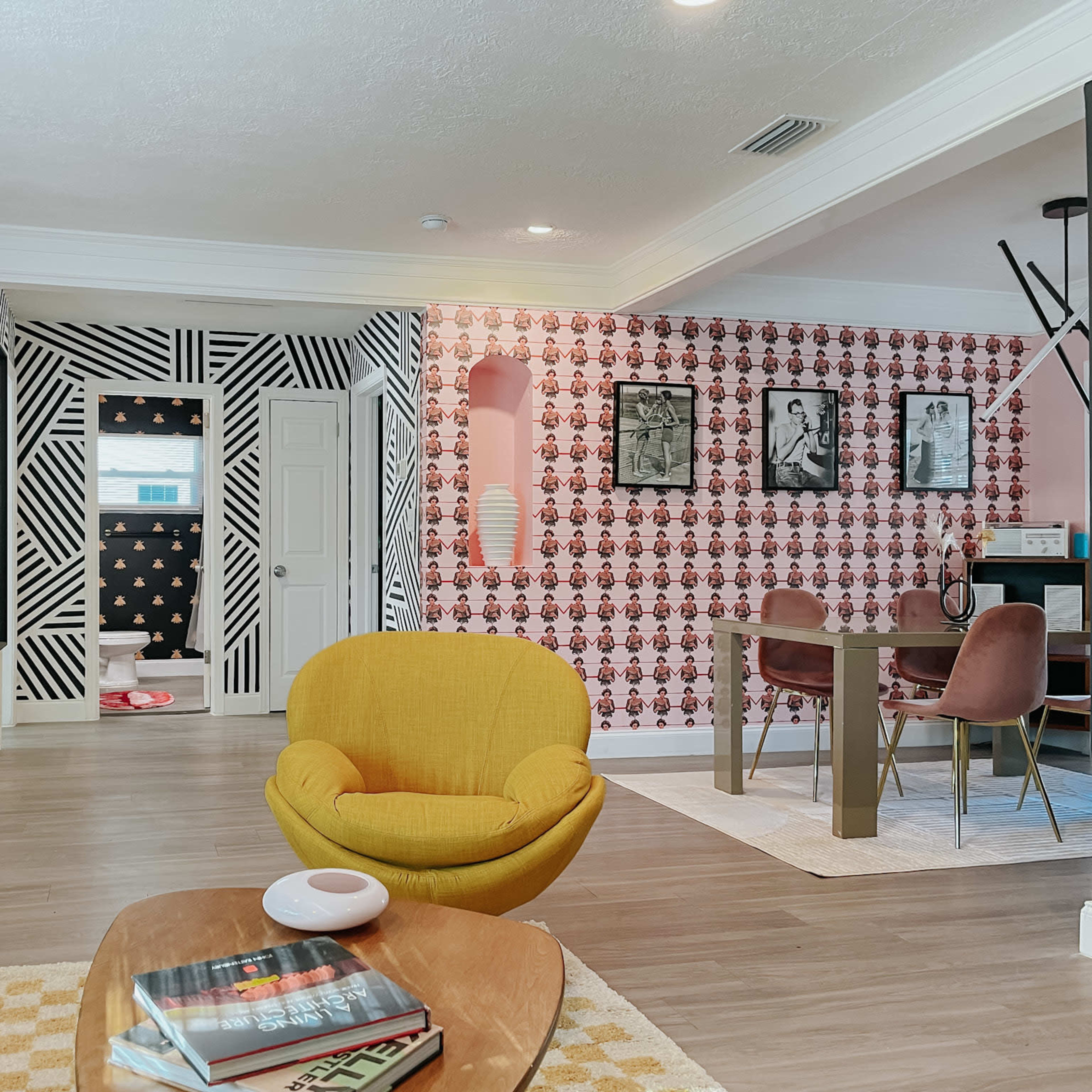 A brightly decorated living space featuring colorful wallpaper, a yellow armchair, a wooden coffee table stacked with books, and a dining area with a dark table against contrasting wall patterns.