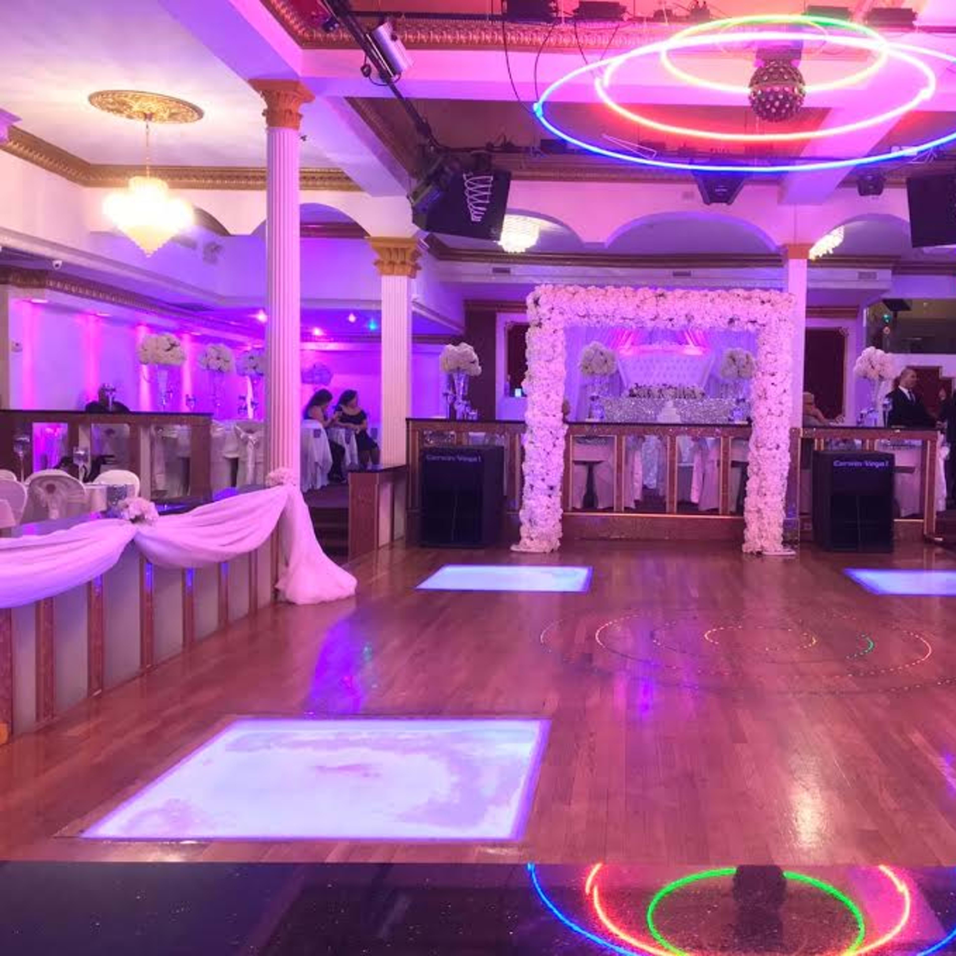 A decorated event space with a wooden dance floor, floral arch at the front, and colorful lighting.