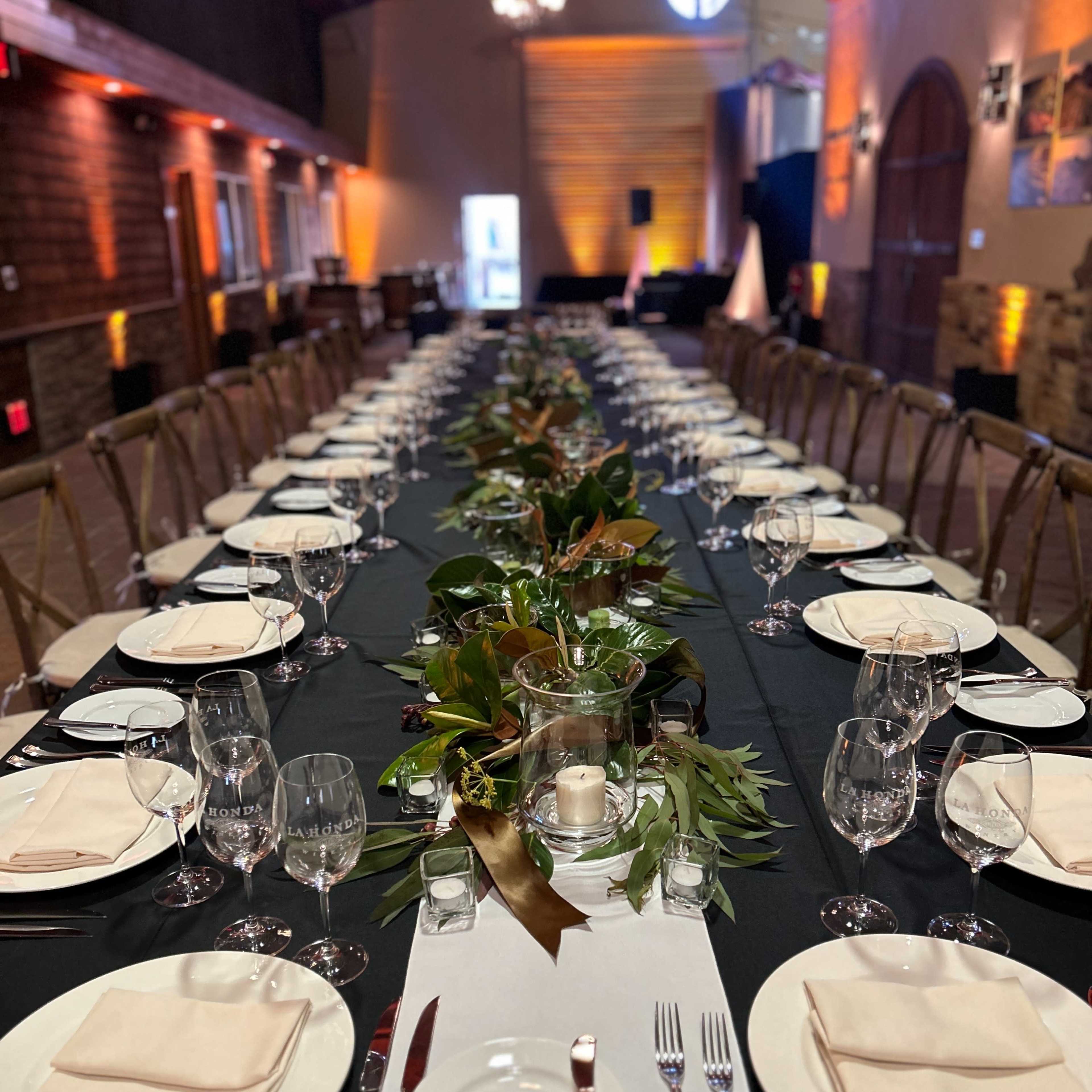 A long, elegantly set dining table is arranged with white plates, glassware, and cloth napkins, featuring a central greenery centerpiece in a spacious venue.