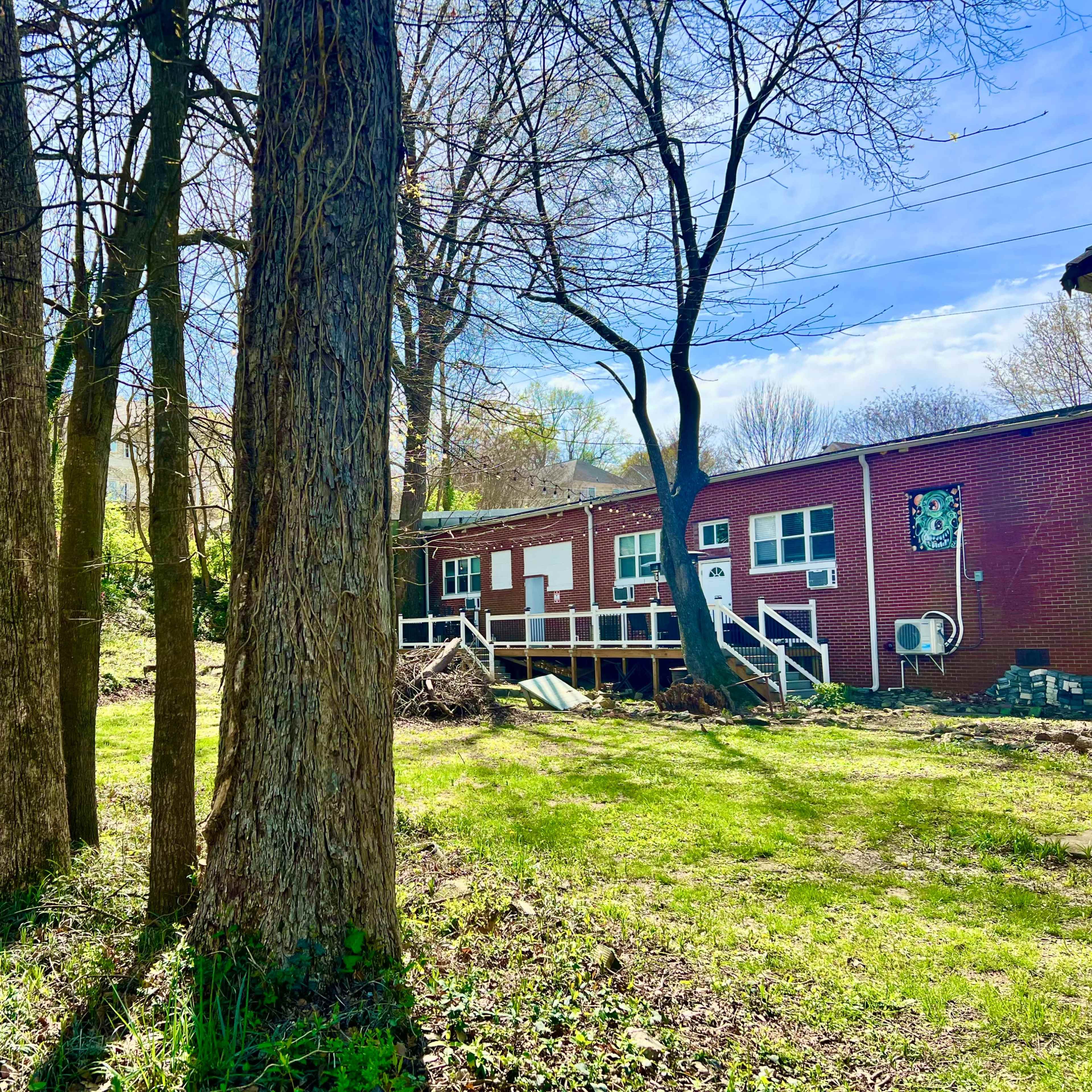 A red brick building with a wooden deck is partially hidden by tall trees in a grassy area.