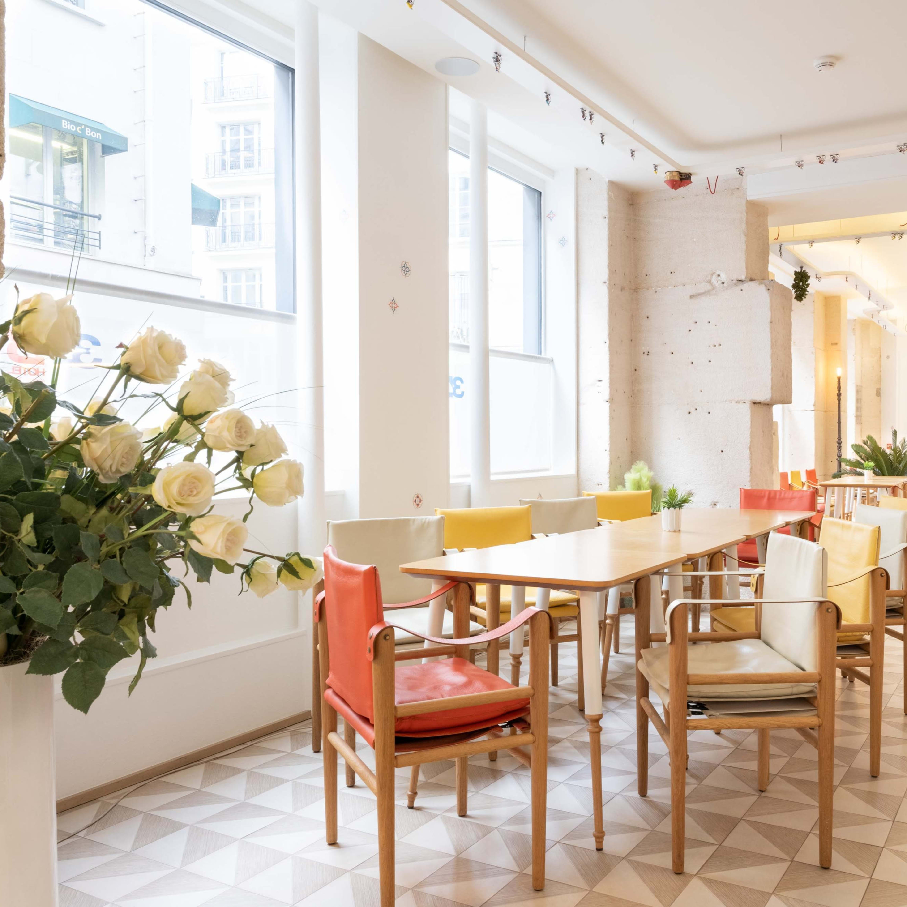 A bright, modern cafe with a series of wooden tables and colorful chairs arranged under large windows.