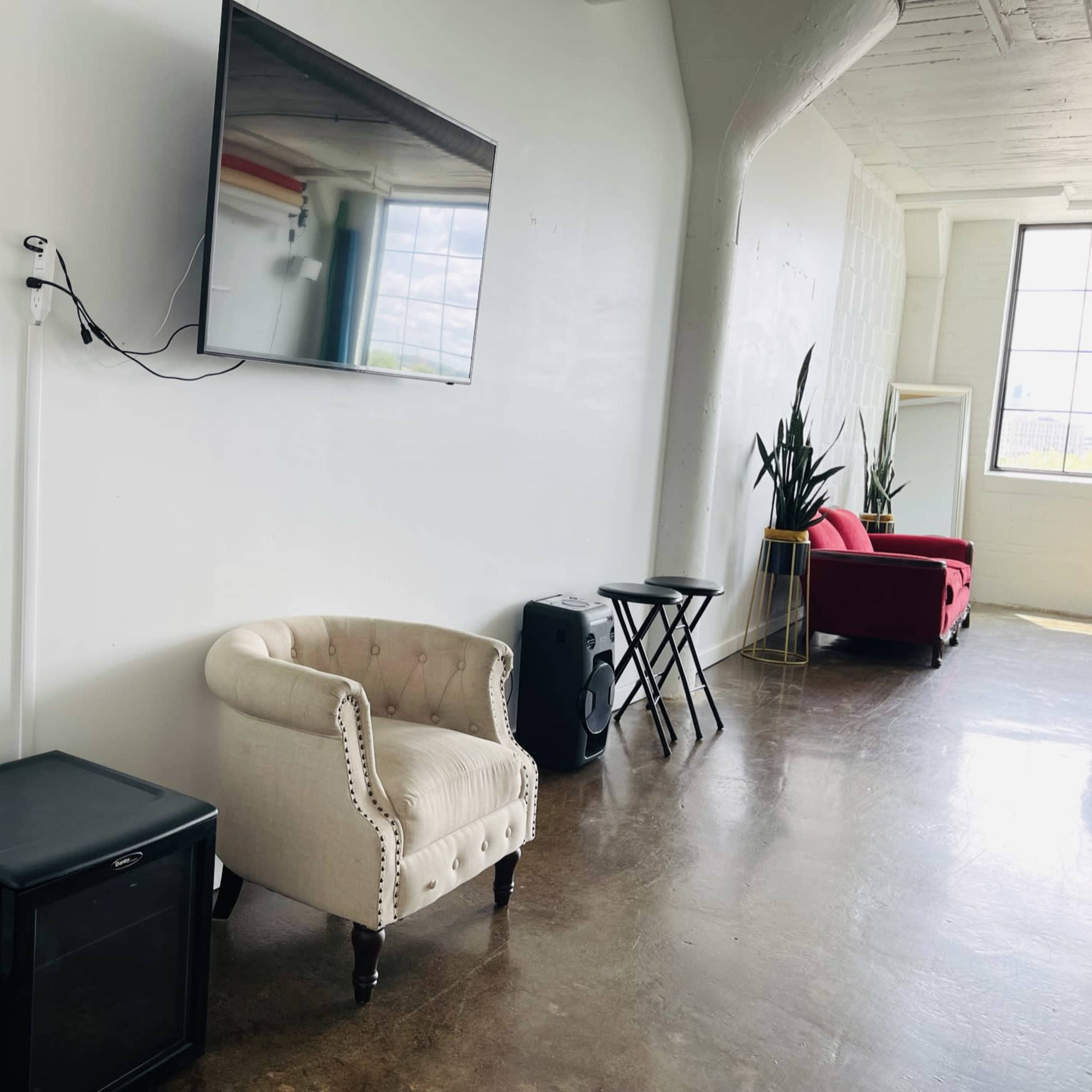 A room with a wall-mounted television, a black mini fridge, a cream-colored armchair, a small side table, and two red chairs, with a large window in the background.