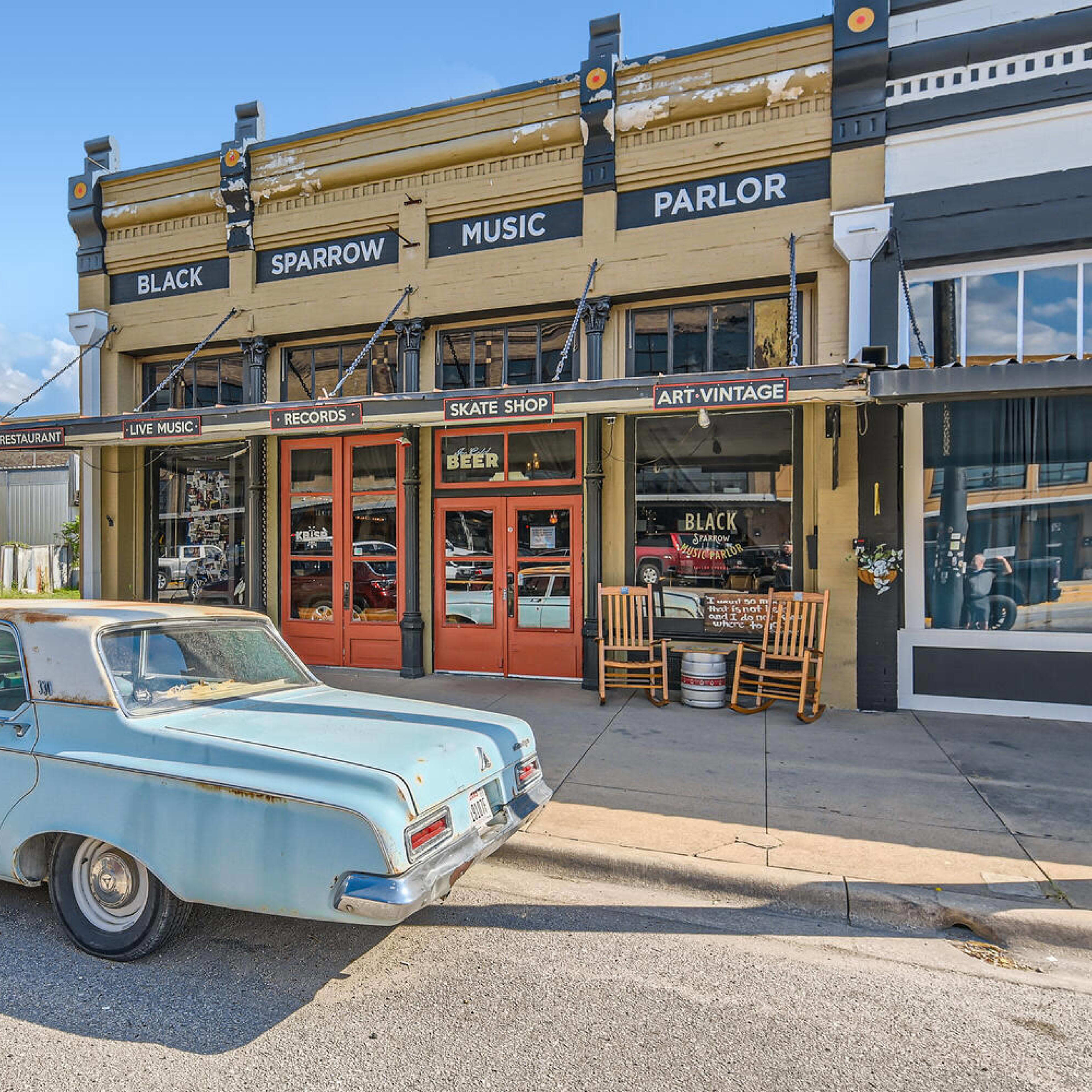 A light blue vintage car is parked in front of a music parlor labeled "Black Sparrow" on a sunny street with a mix of shops.