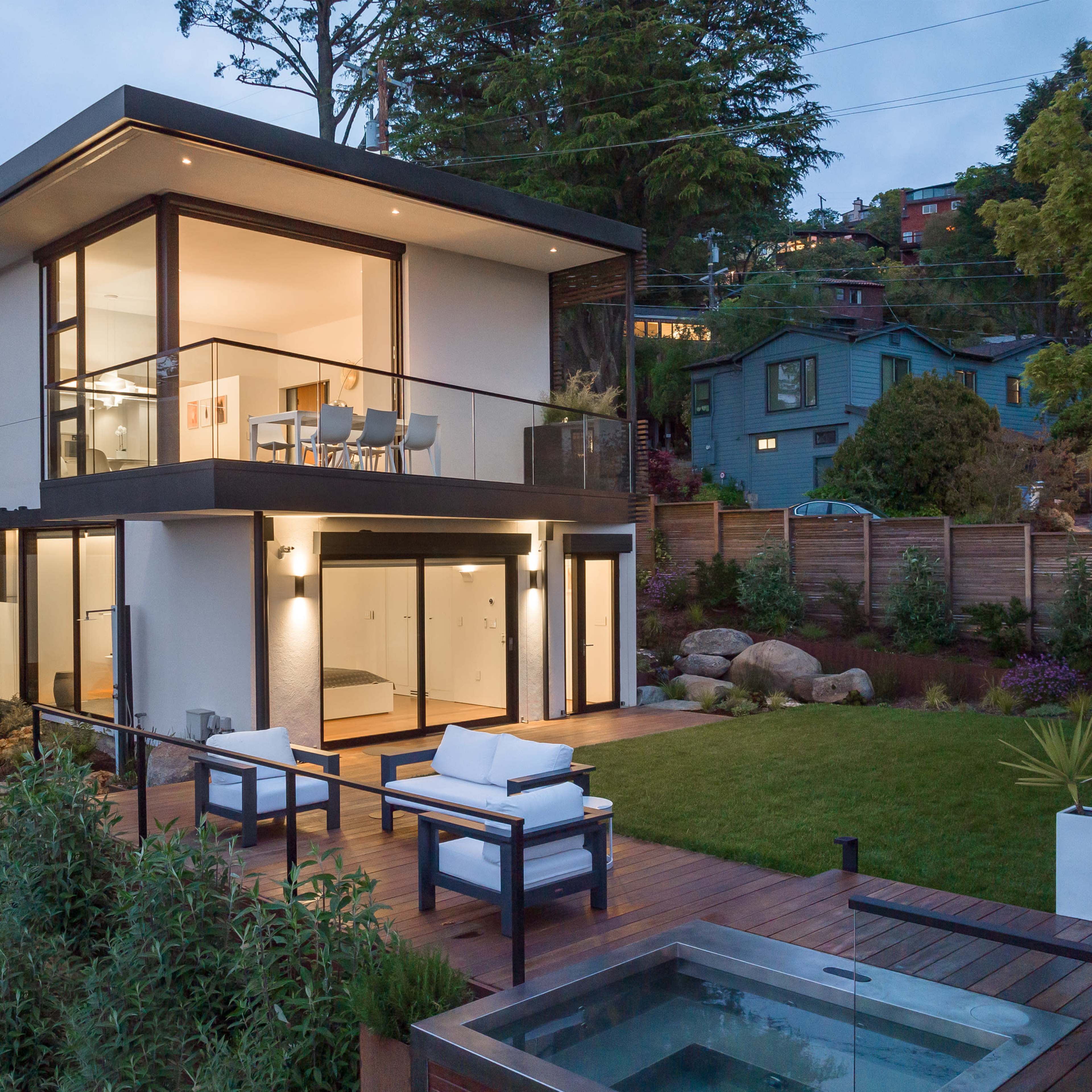 A modern two-story house features large glass windows, a patio with outdoor seating, and a landscaped yard with a hot tub.