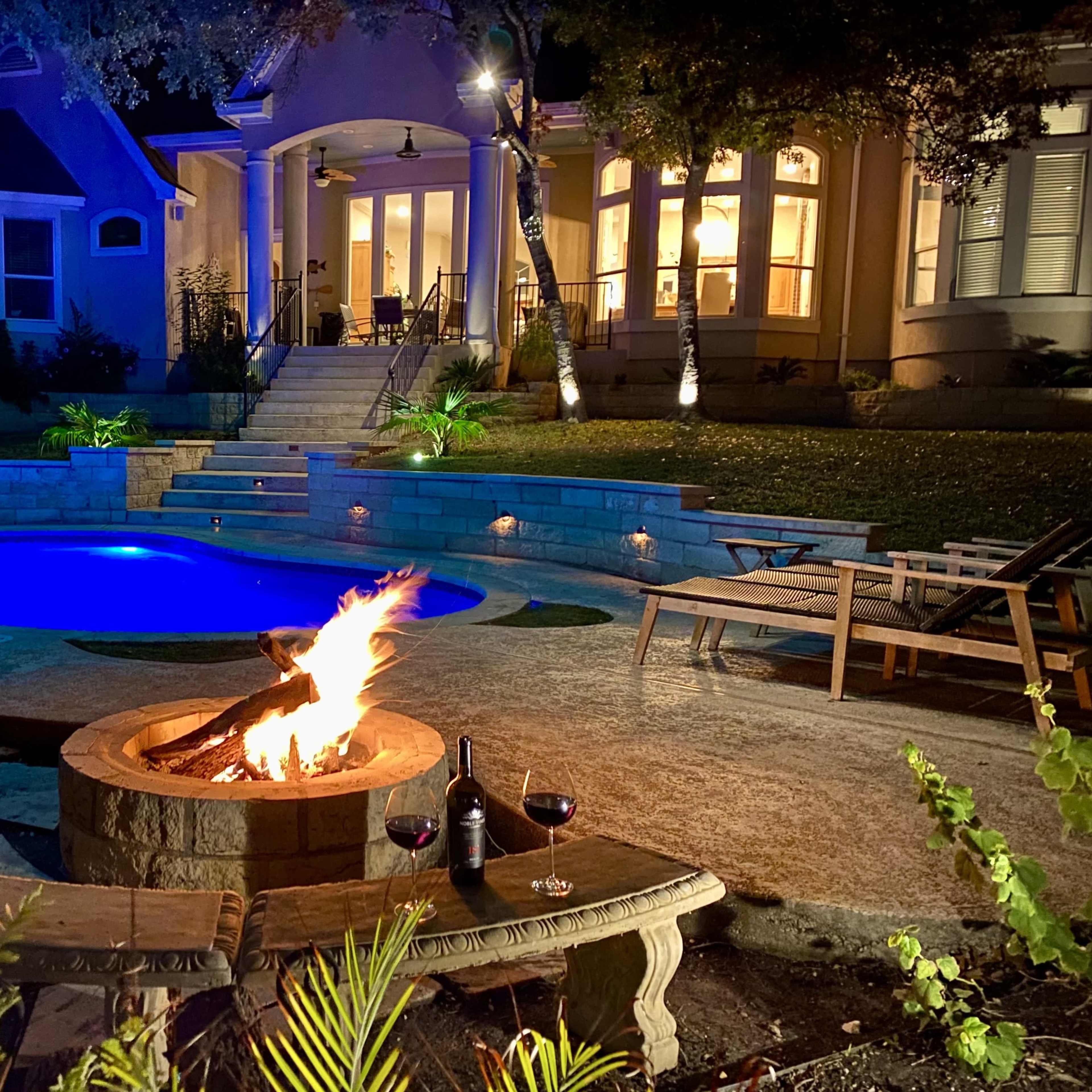 A fire pit surrounded by stone seating is illuminated by soft lighting in a backyard with a pool and a house in the background.