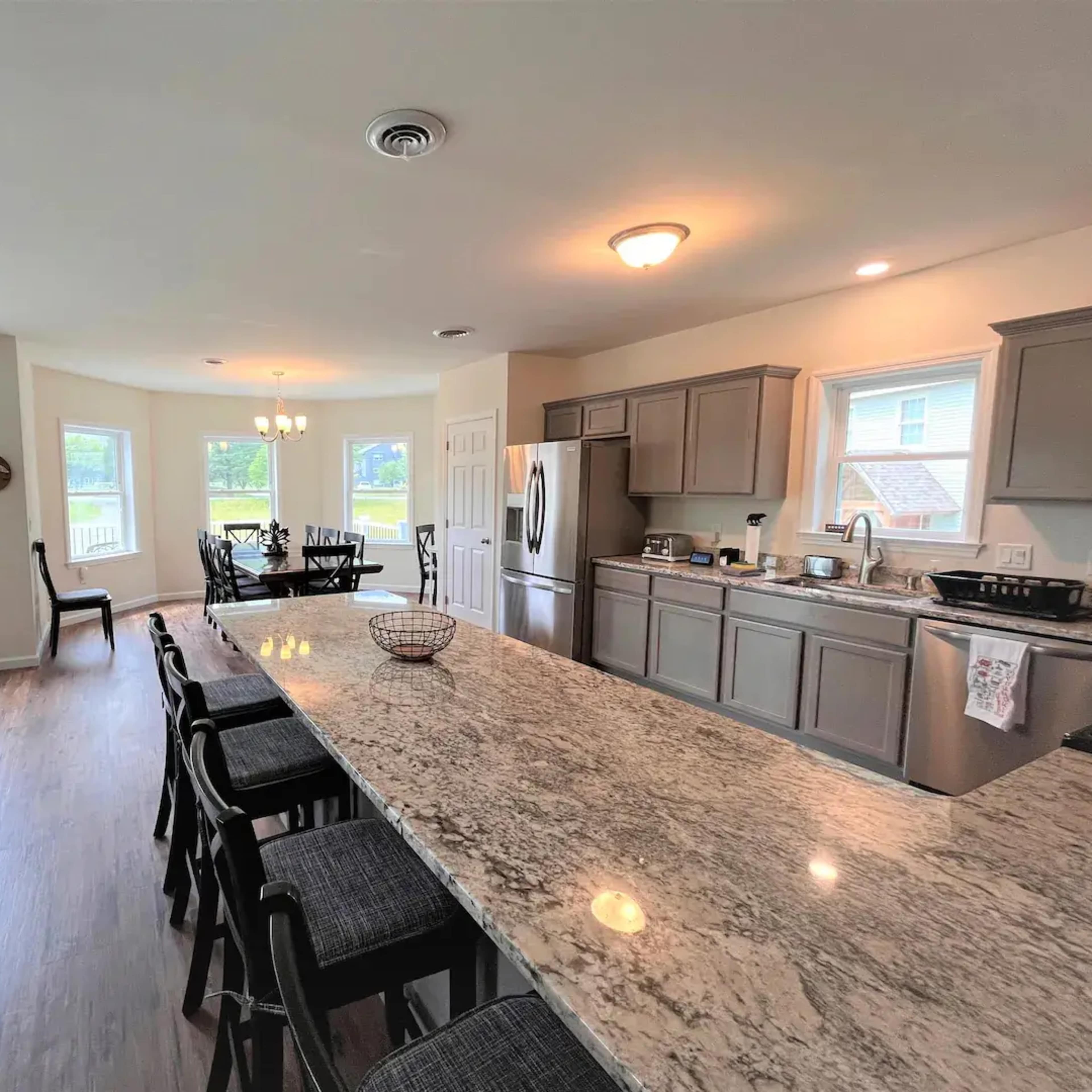 A spacious kitchen with gray cabinets, a large granite island with bar seating, and windows overlooking a green outdoor area.