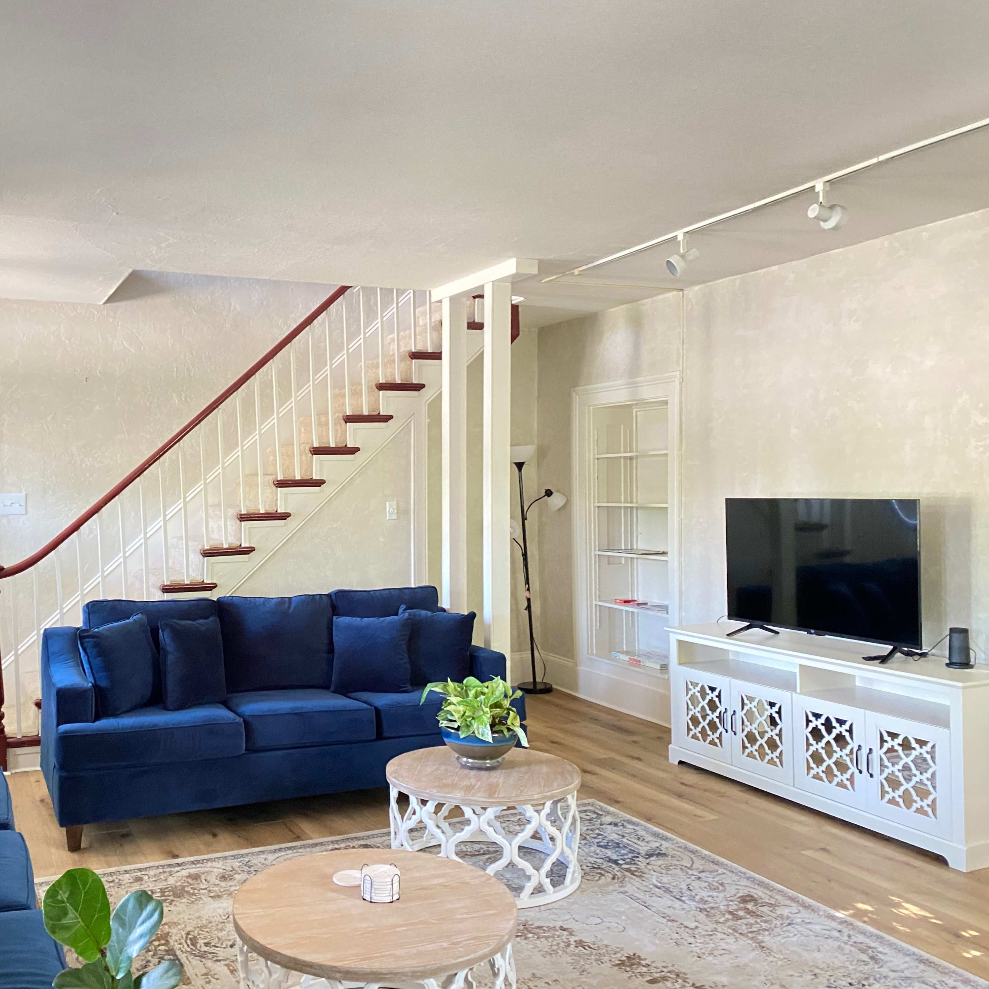 A living room with a blue sofa set, a round coffee table, a television on a white cabinet, and a staircase in the background.