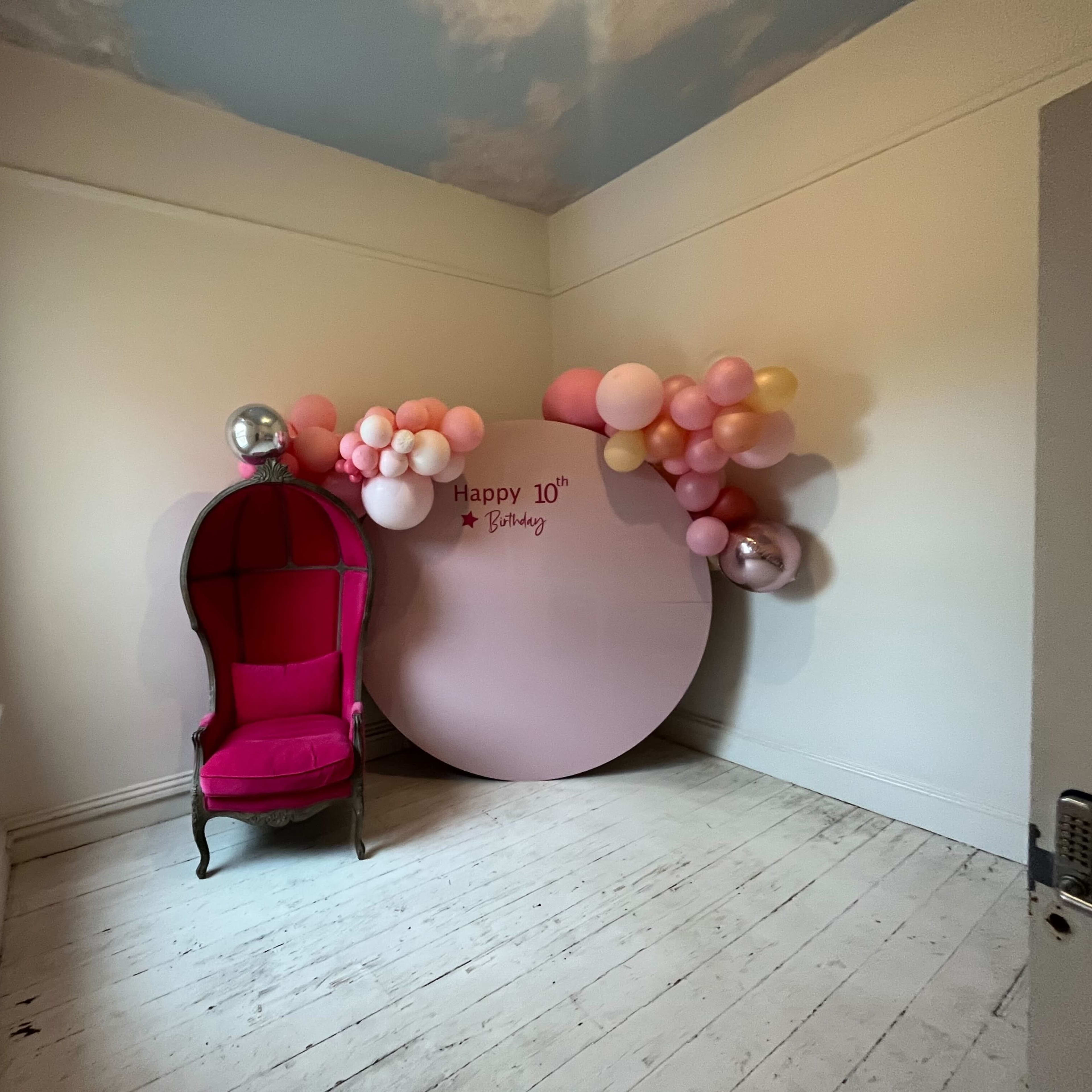 A corner of a room featuring a pink chair and a decorated wall with a round sign that says "Happy 10th Birthday," surrounded by an arrangement of pink and silver balloons.
