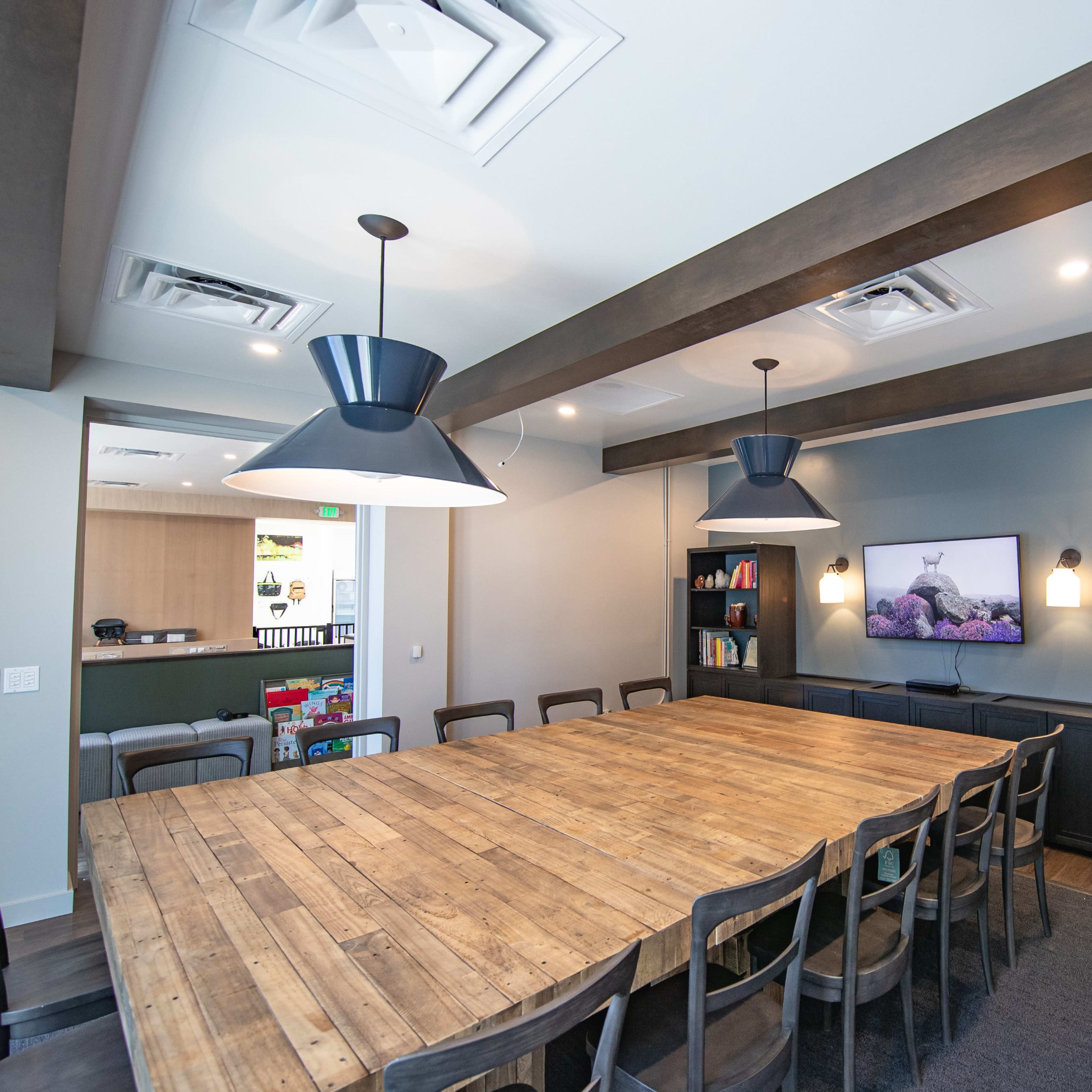 A large wooden table with black chairs is set in a conference room featuring overhead pendant lights and a TV mounted on a blue wall.