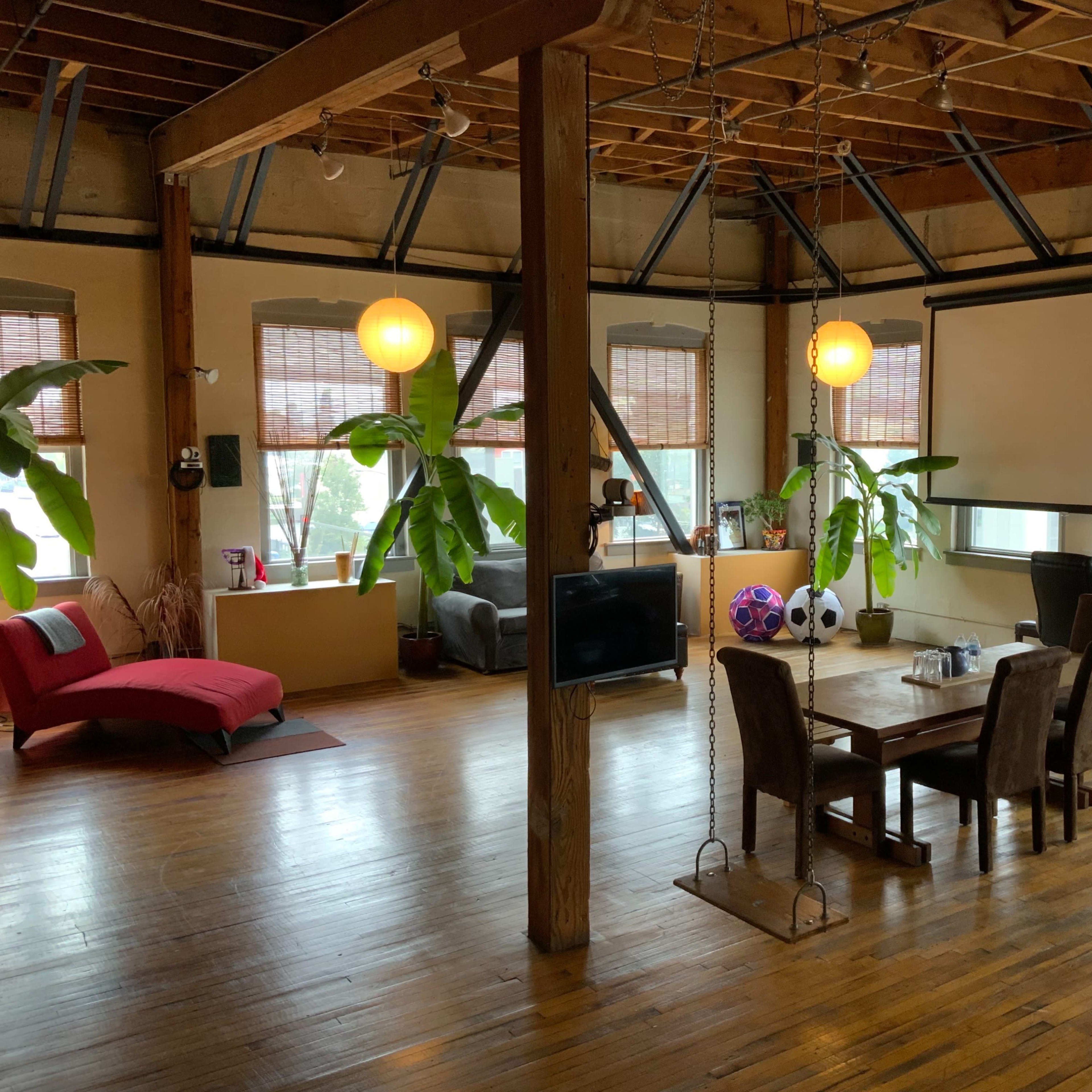 A spacious loft interior featuring wooden floors, large windows, a dining table, and seating areas with plants and decorative lighting.