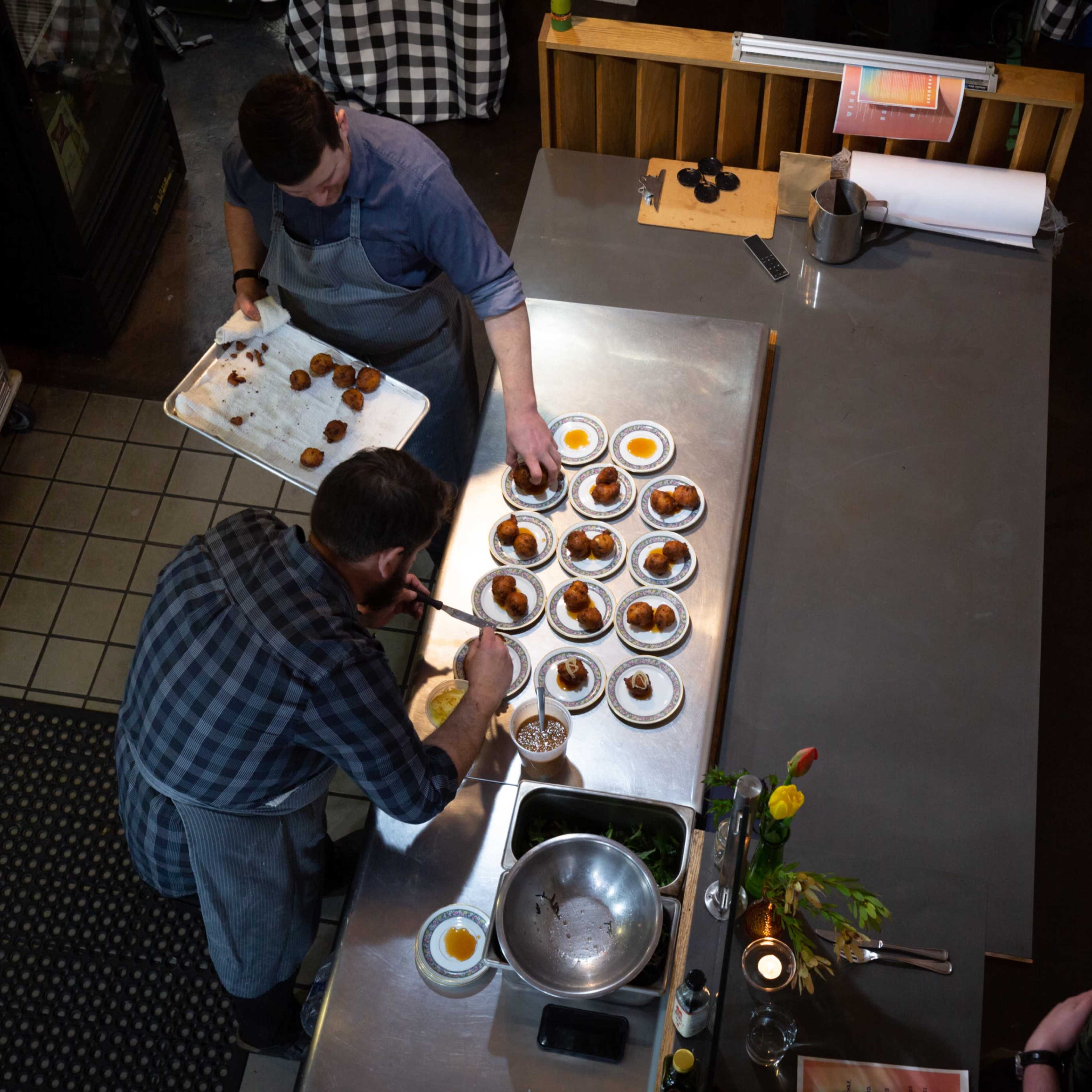 Two chefs are plating dishes on a stainless steel counter in a kitchen.