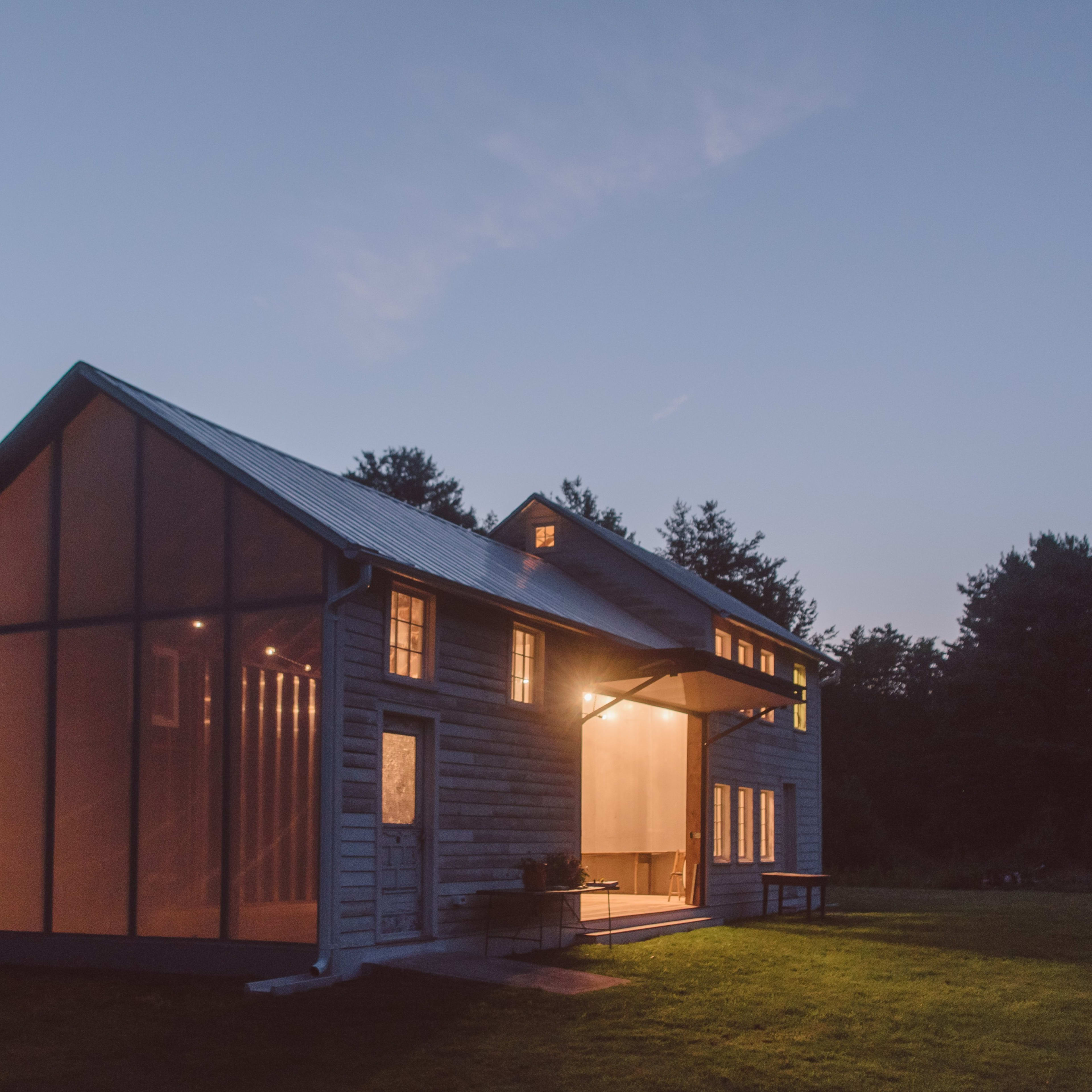 A modern, two-story house with large windows and a screened porch is illuminated at dusk, surrounded by trees.