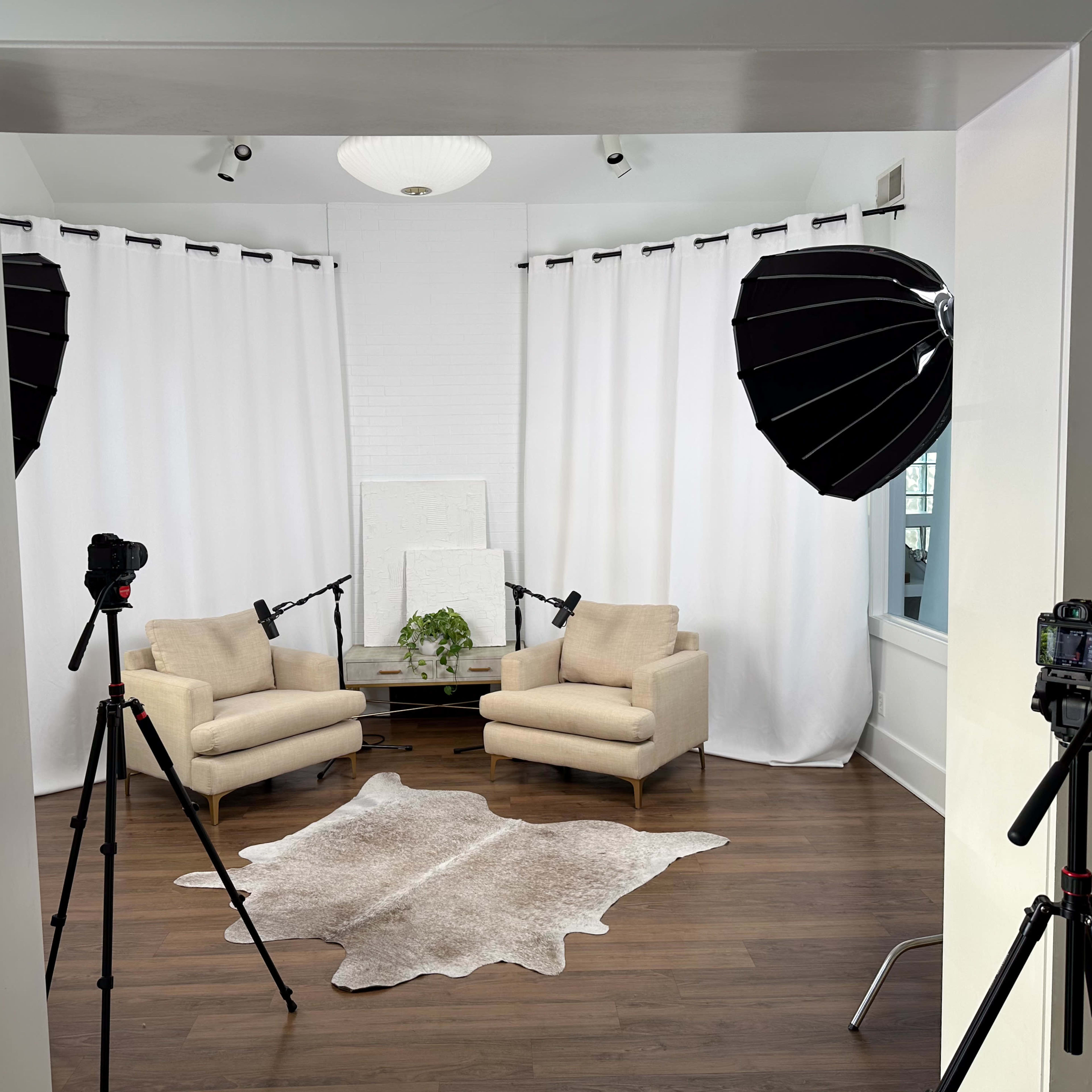 A well-lit studio setup features two beige chairs, a coffee table, and soft lighting equipment arranged in front of white curtains.