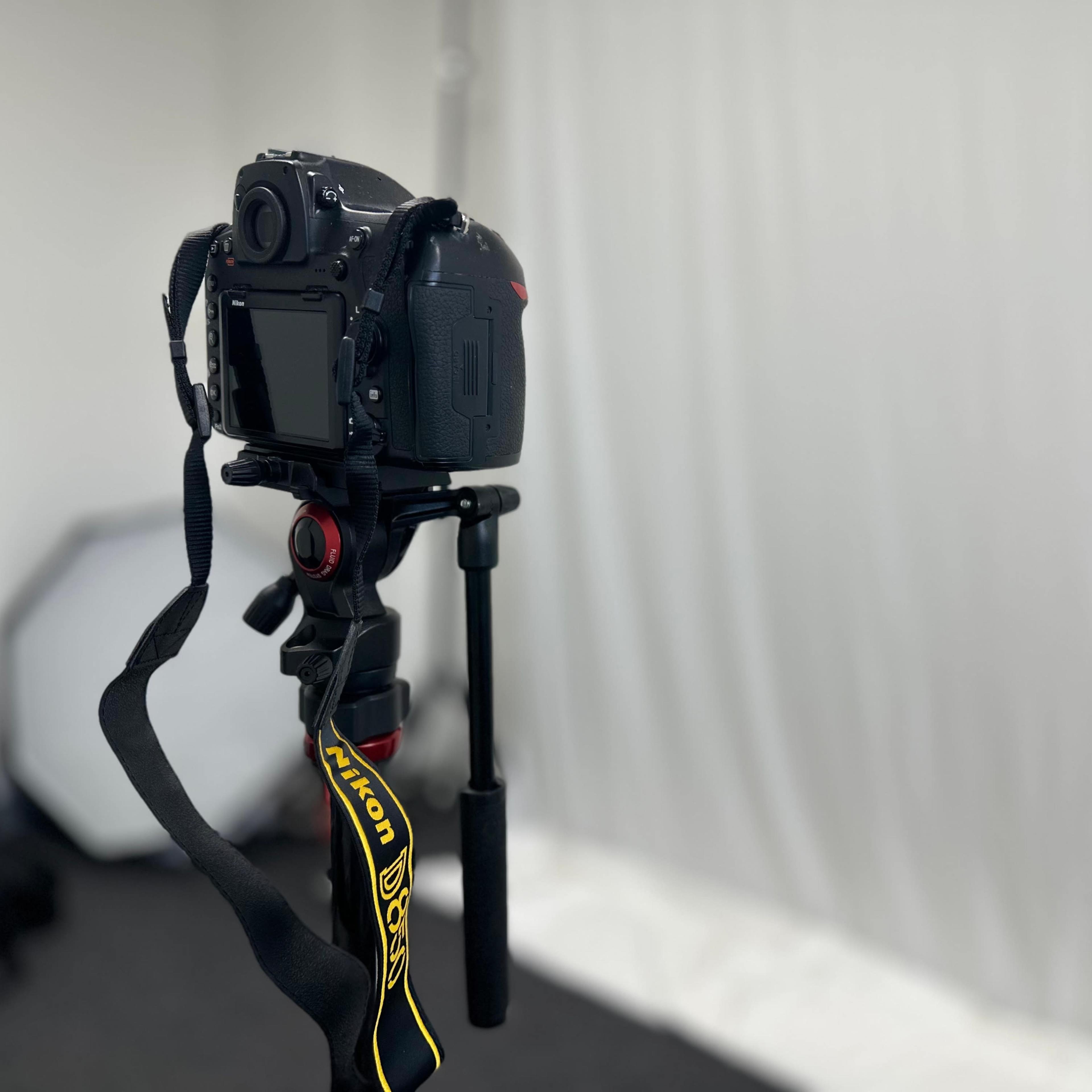 A Nikon camera is mounted on a tripod, facing a plain white backdrop in a studio setting.