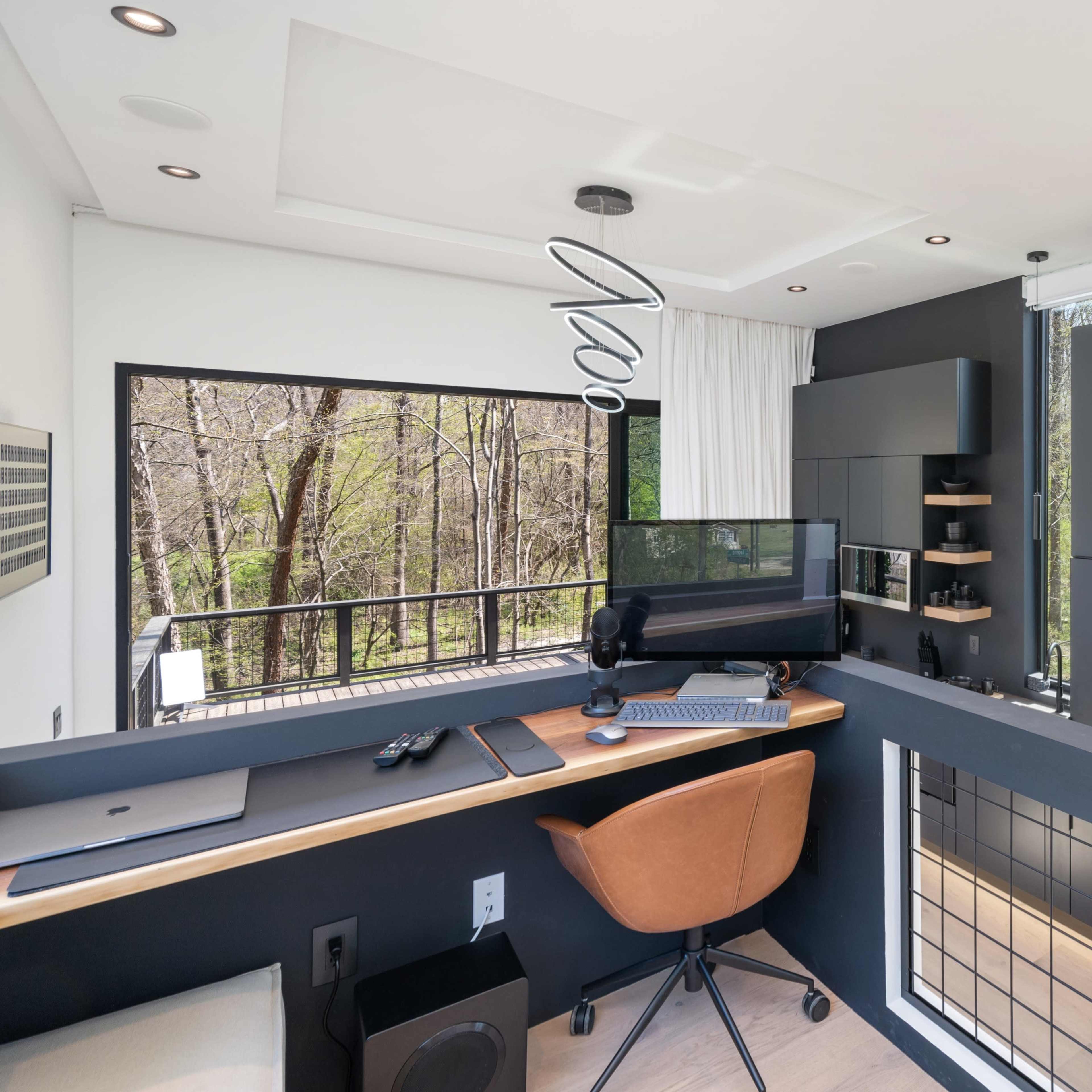 A modern workspace features a desk with a computer, shelves, and a view of trees through large windows.