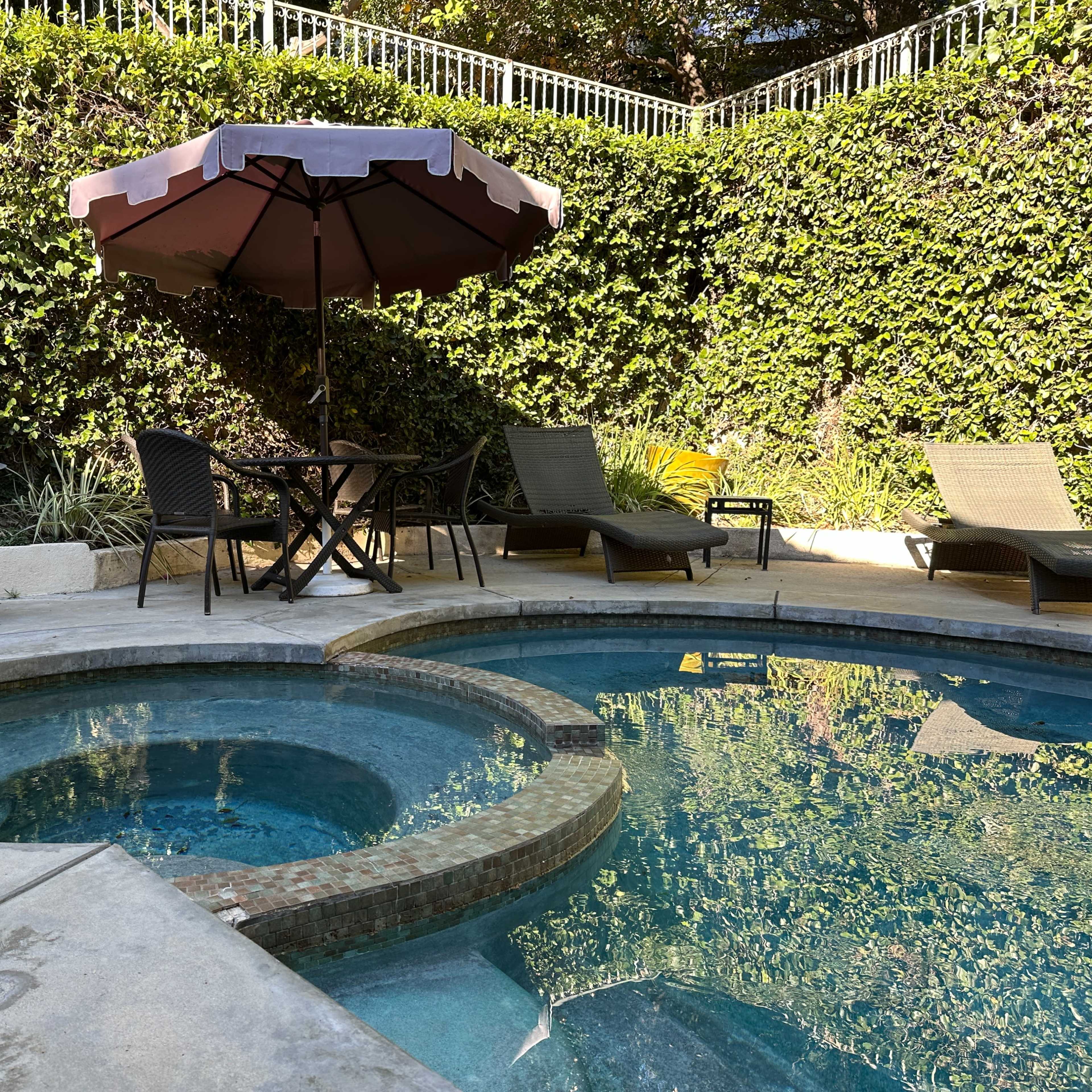 A small outdoor pool area features a hot tub, a table with an umbrella, and several lounge chairs surrounded by greenery.