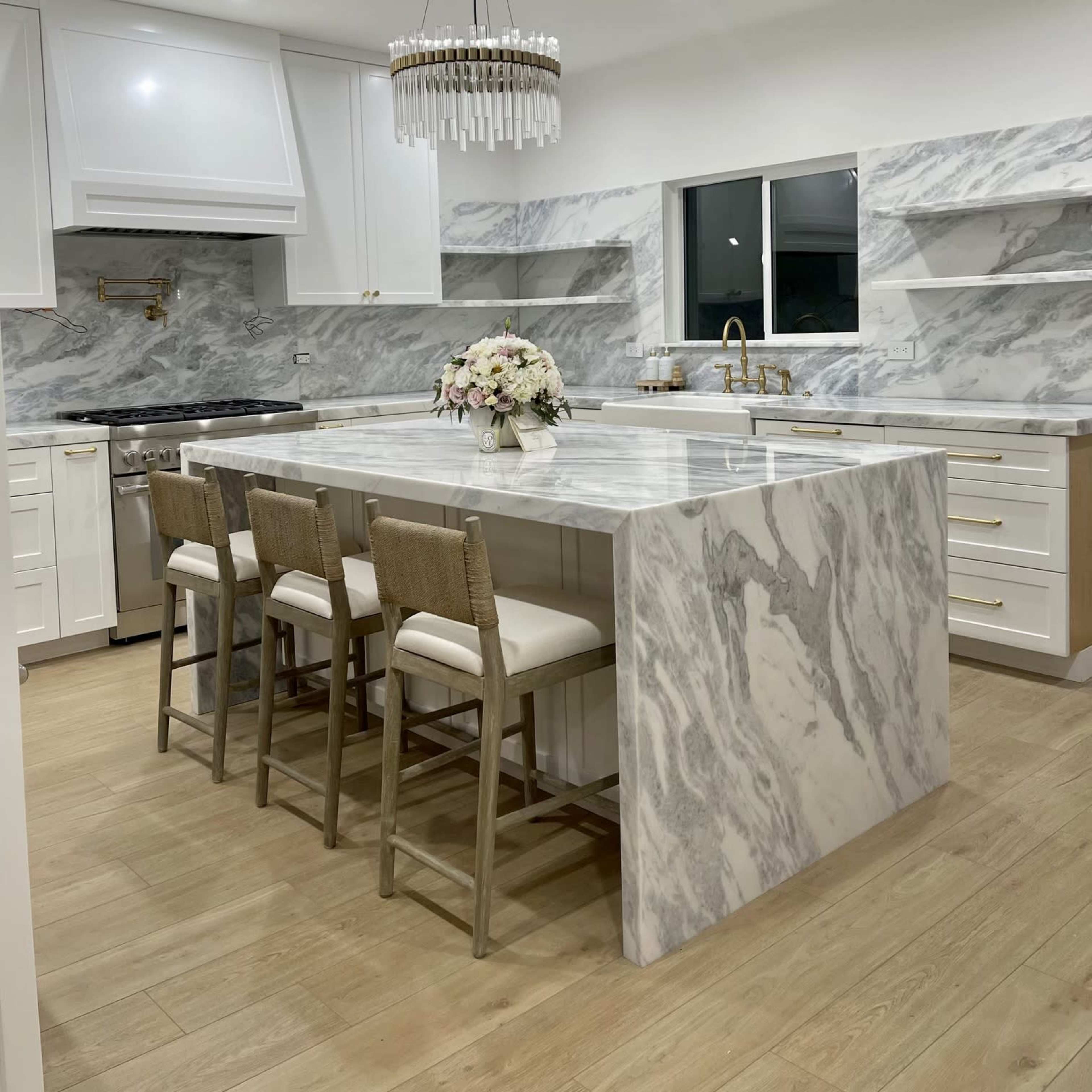 A modern kitchen features a large marble island with seating, white cabinetry, and a chandelier above.