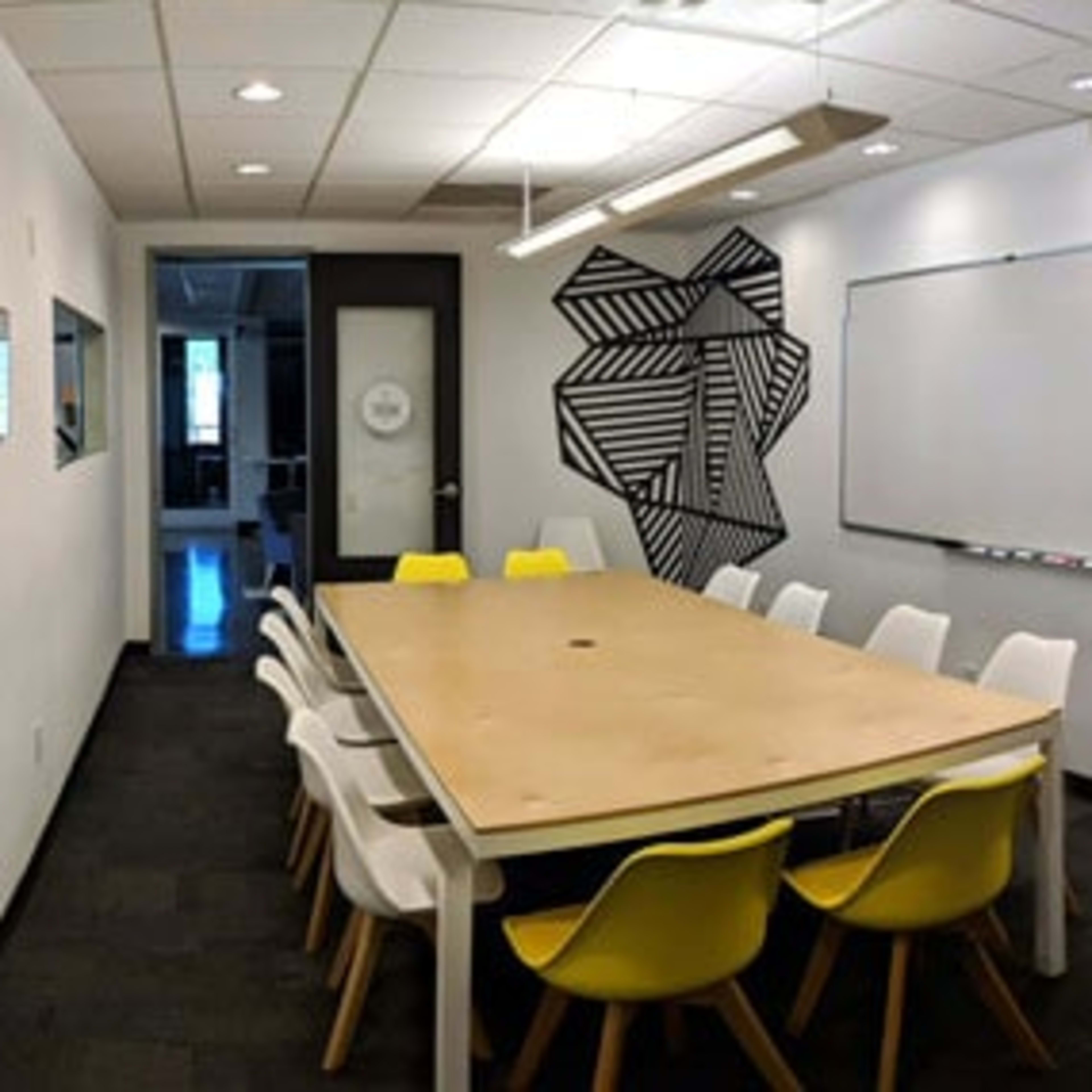 A modern conference room with a large wooden table surrounded by white and yellow chairs, a whiteboard on one wall, and a minimalist plant on a side shelf.