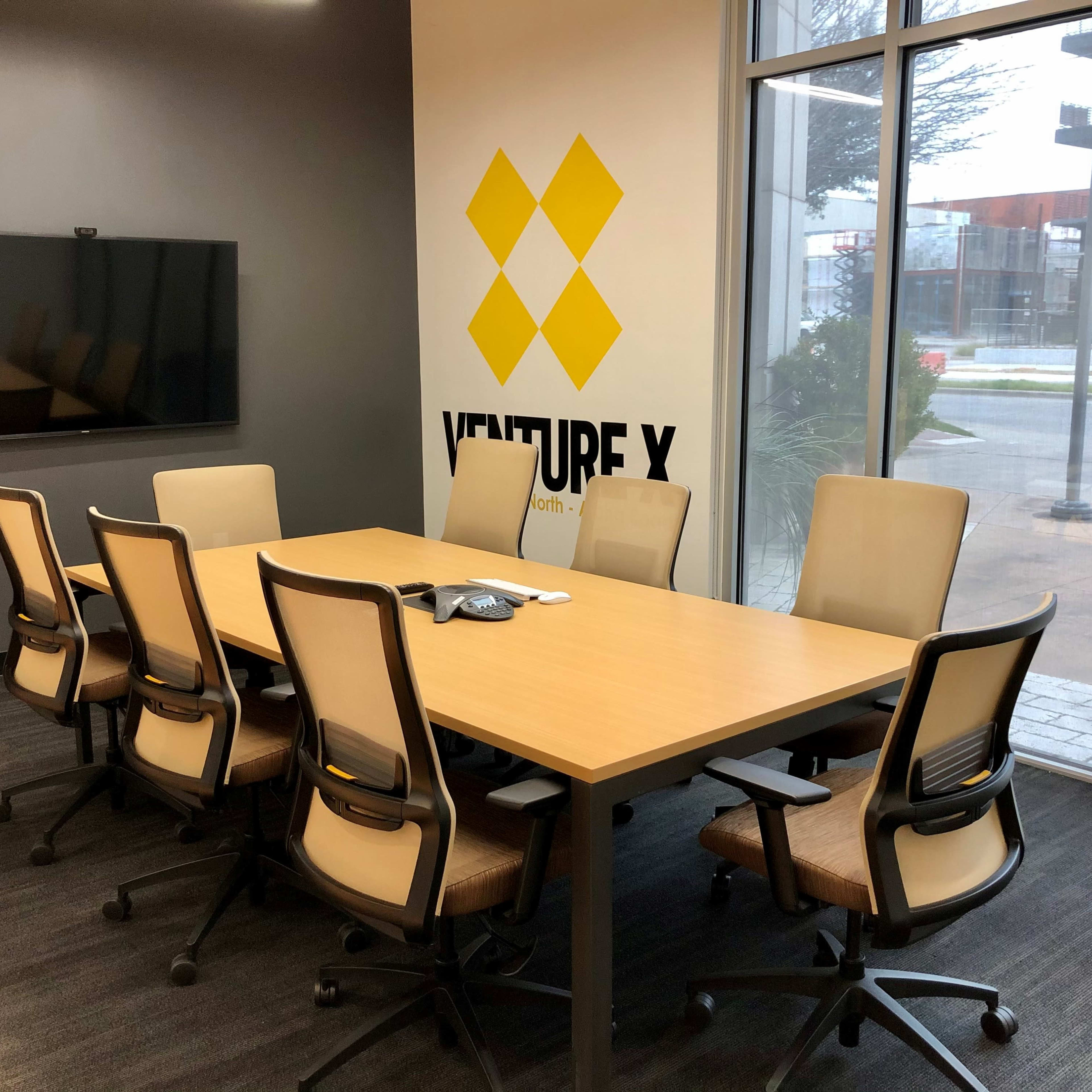 A conference room features a long wooden table surrounded by eight chairs, with a large monitor on the wall and a company logo displayed prominently.