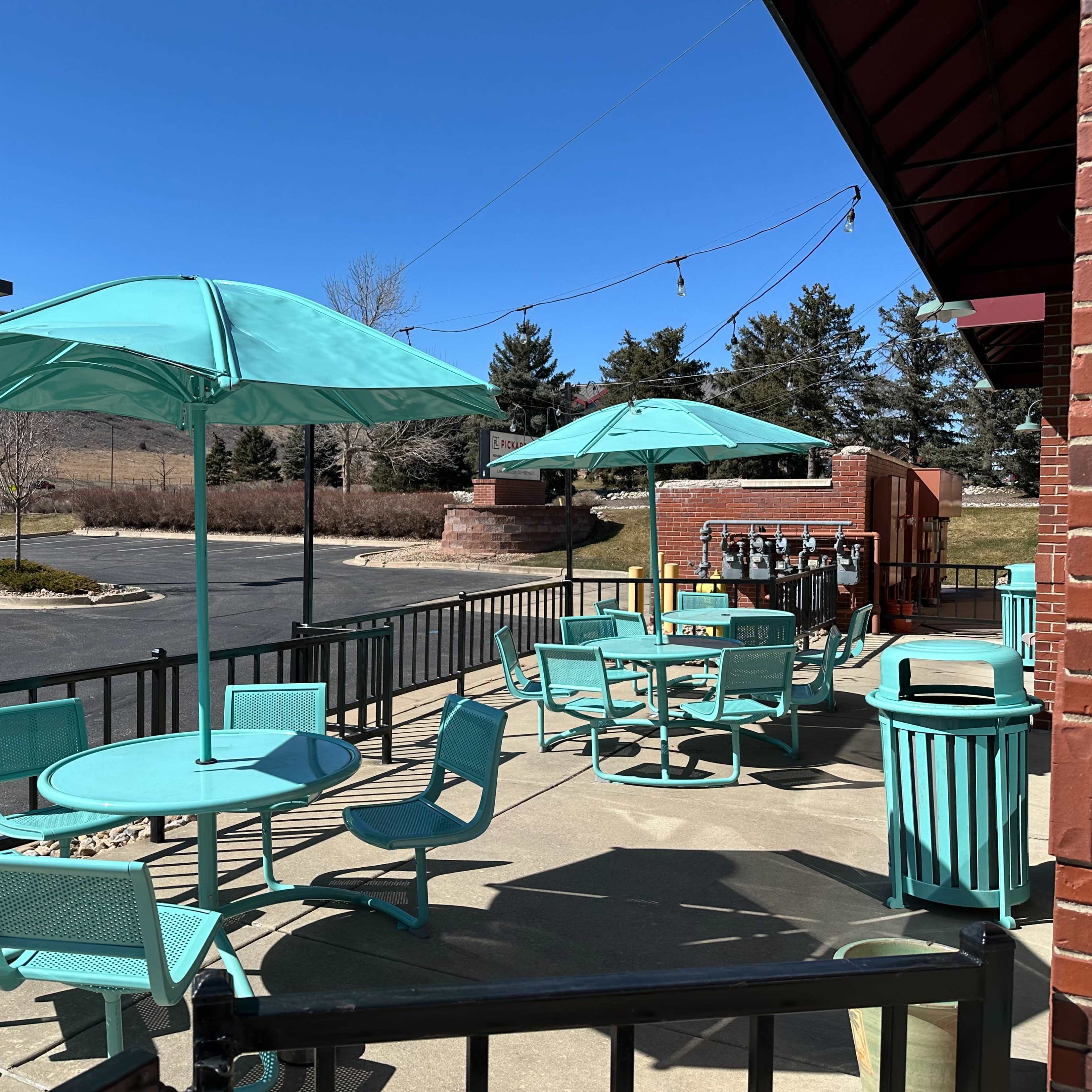 A patio area with turquoise tables and chairs, each with matching umbrellas, situated next to a parking lot and surrounded by grass and trees.