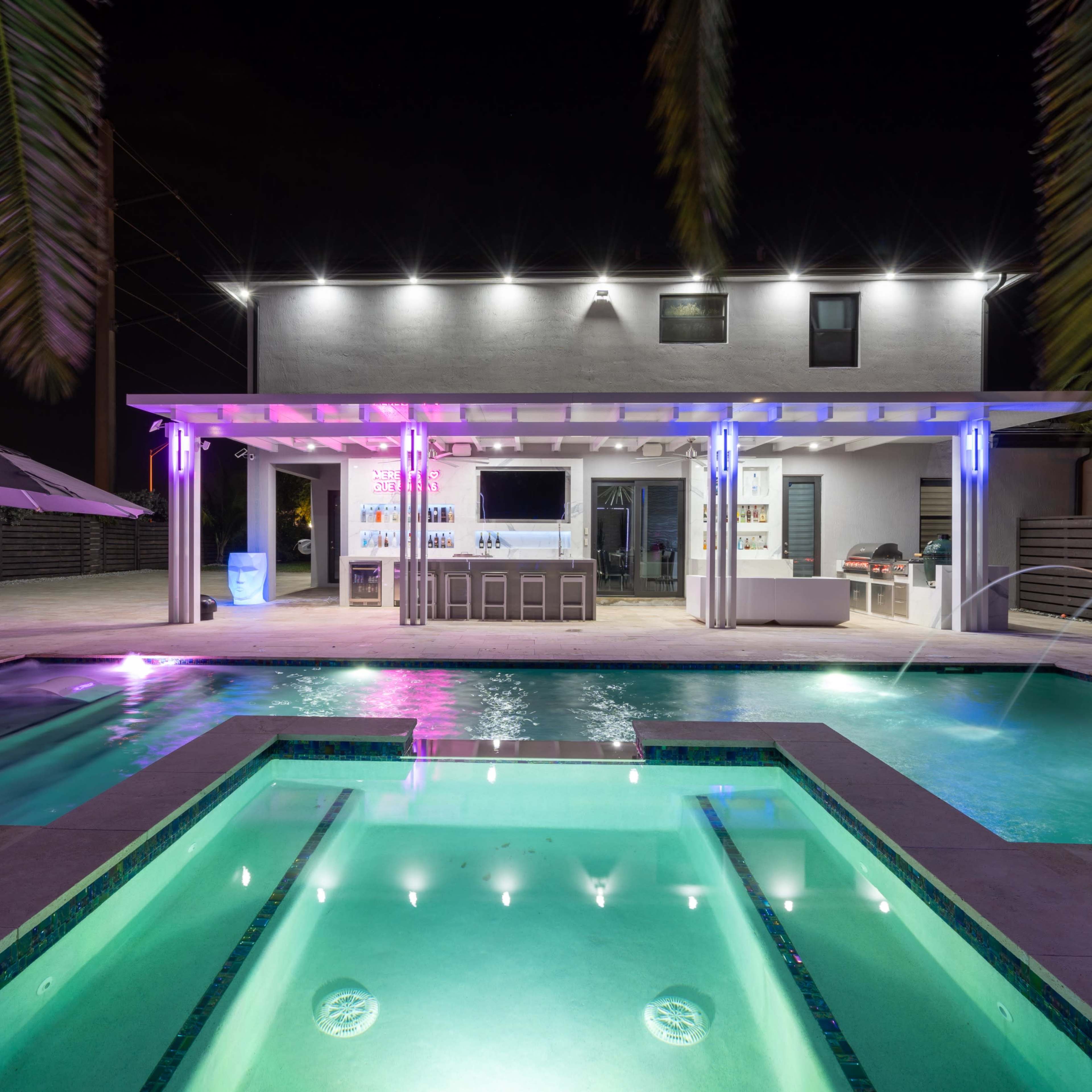 A modern outdoor pool area at night, featuring a rectangular pool with built-in lights, a bar under a pergola, and lounge chairs beside the water.