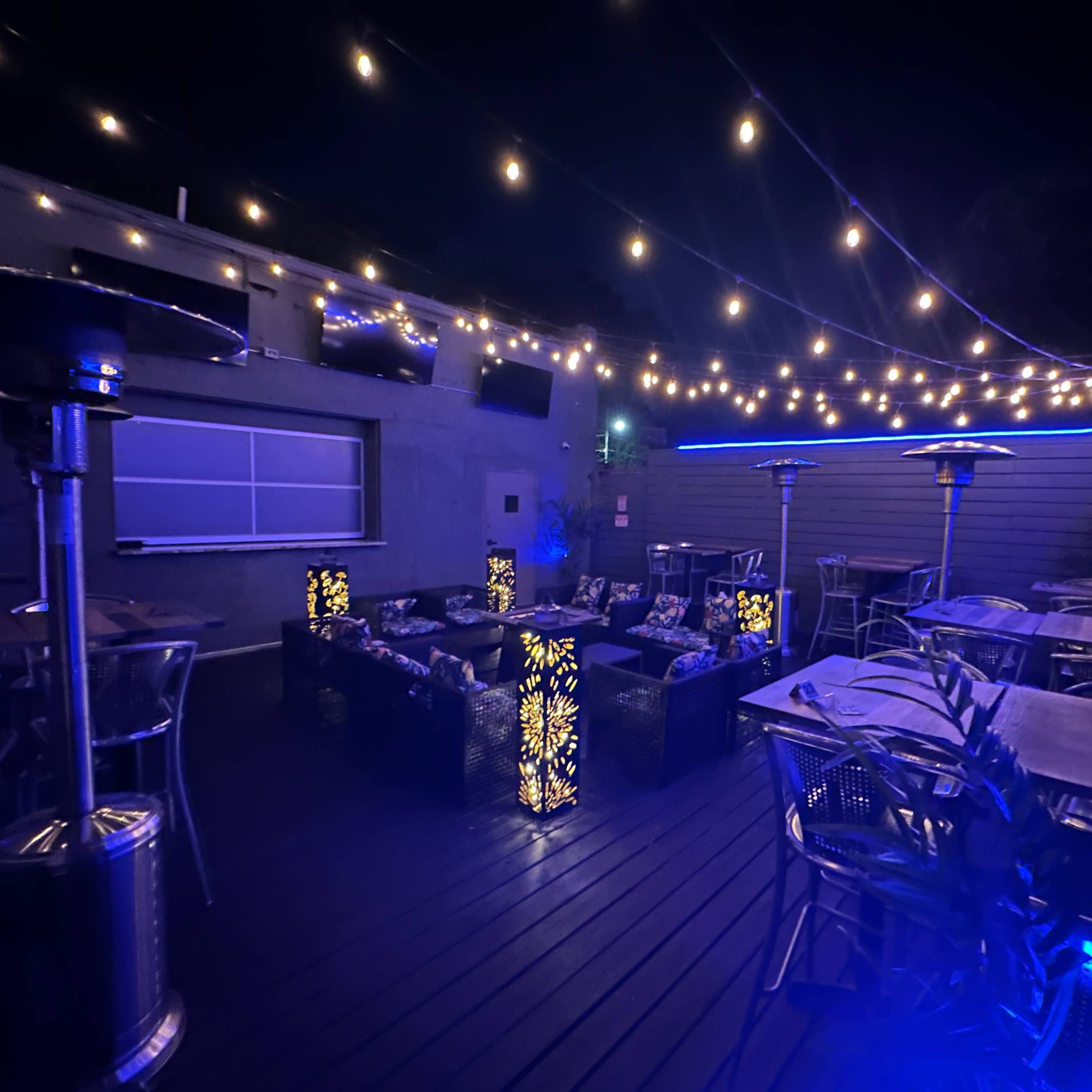 A dimly lit outdoor seating area with string lights and patio heaters, surrounded by tables and chairs.