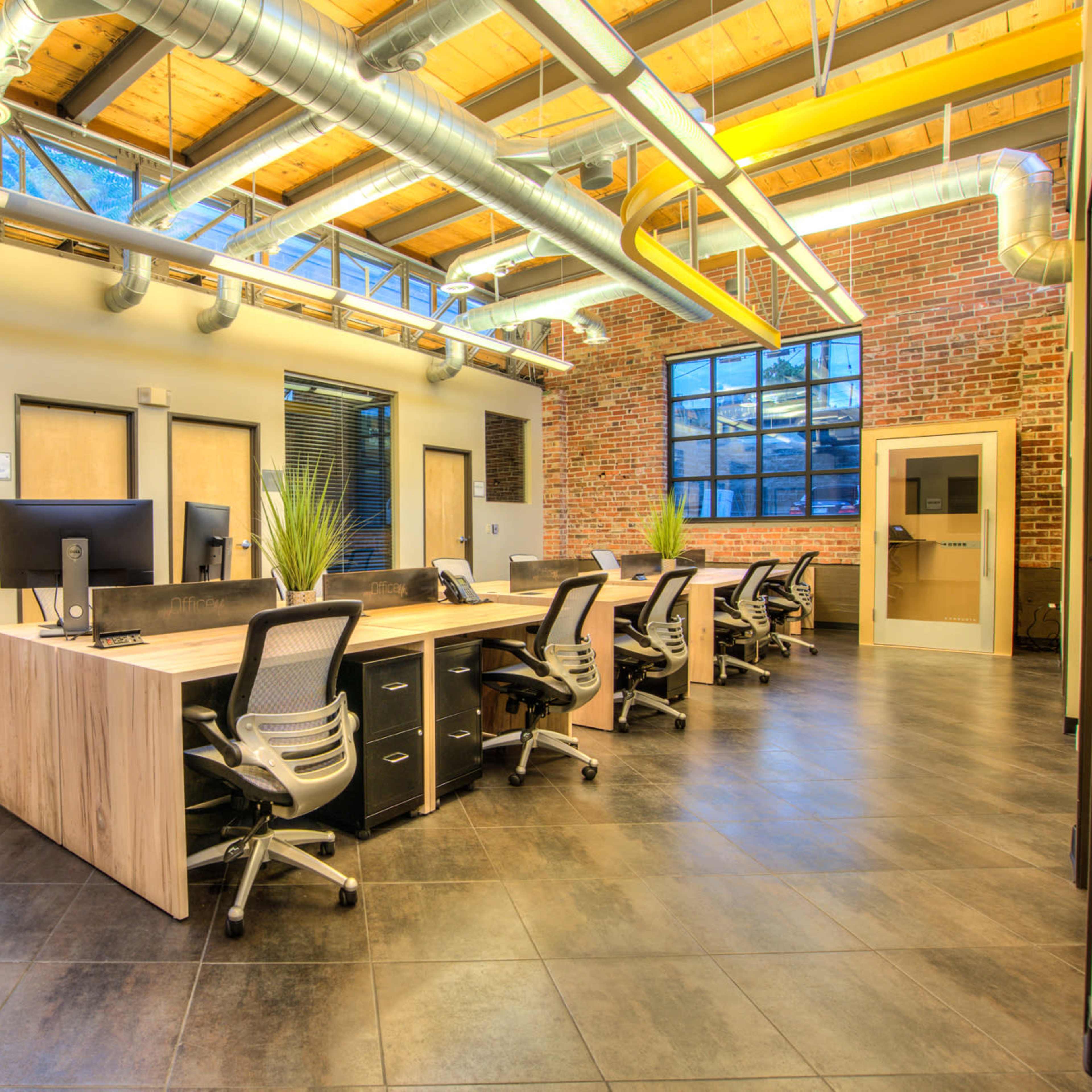 A modern office space with a long central desk surrounded by ergonomic chairs, large windows, and exposed brick walls.