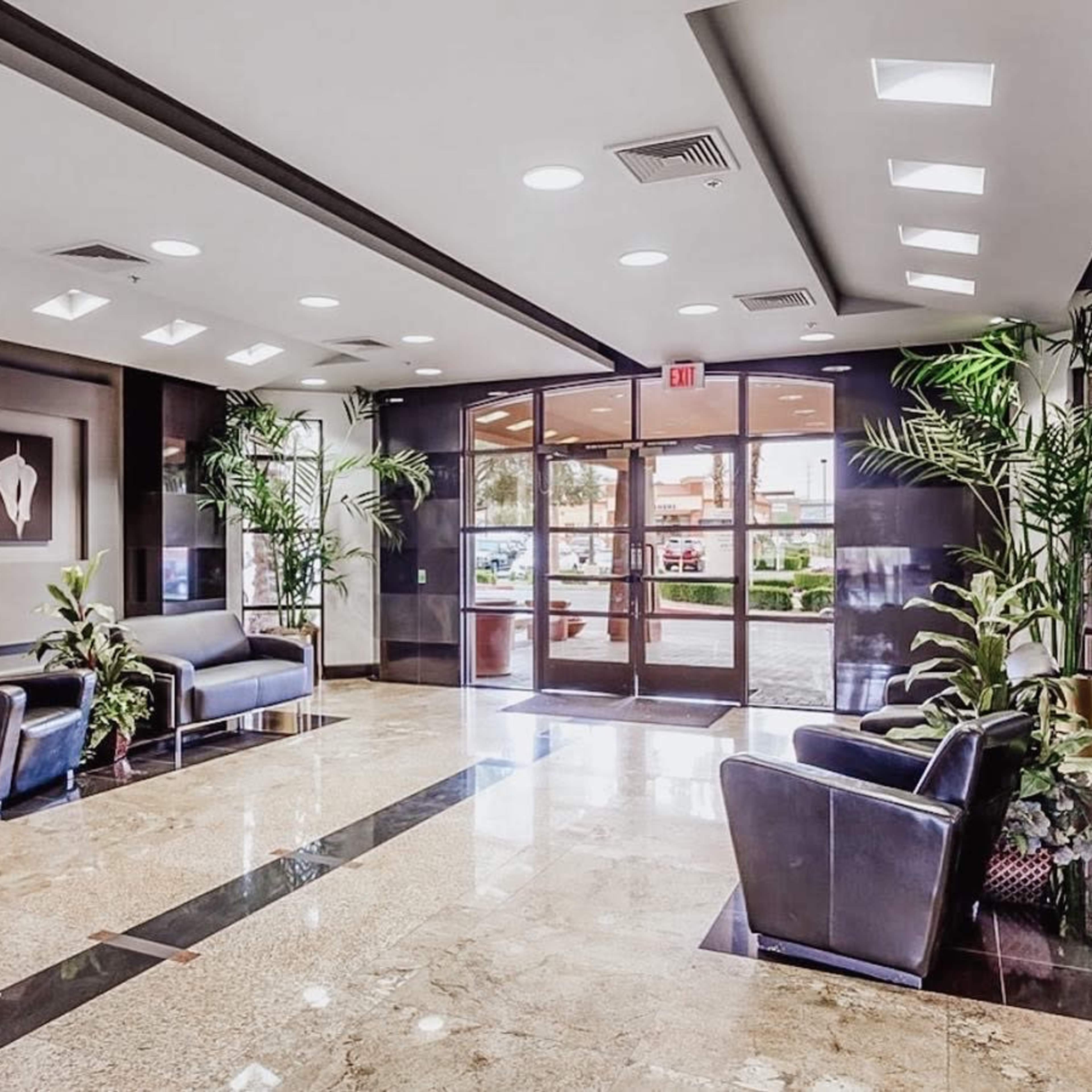 A modern lobby with two black seating areas, potted plants, and large windows allowing natural light.