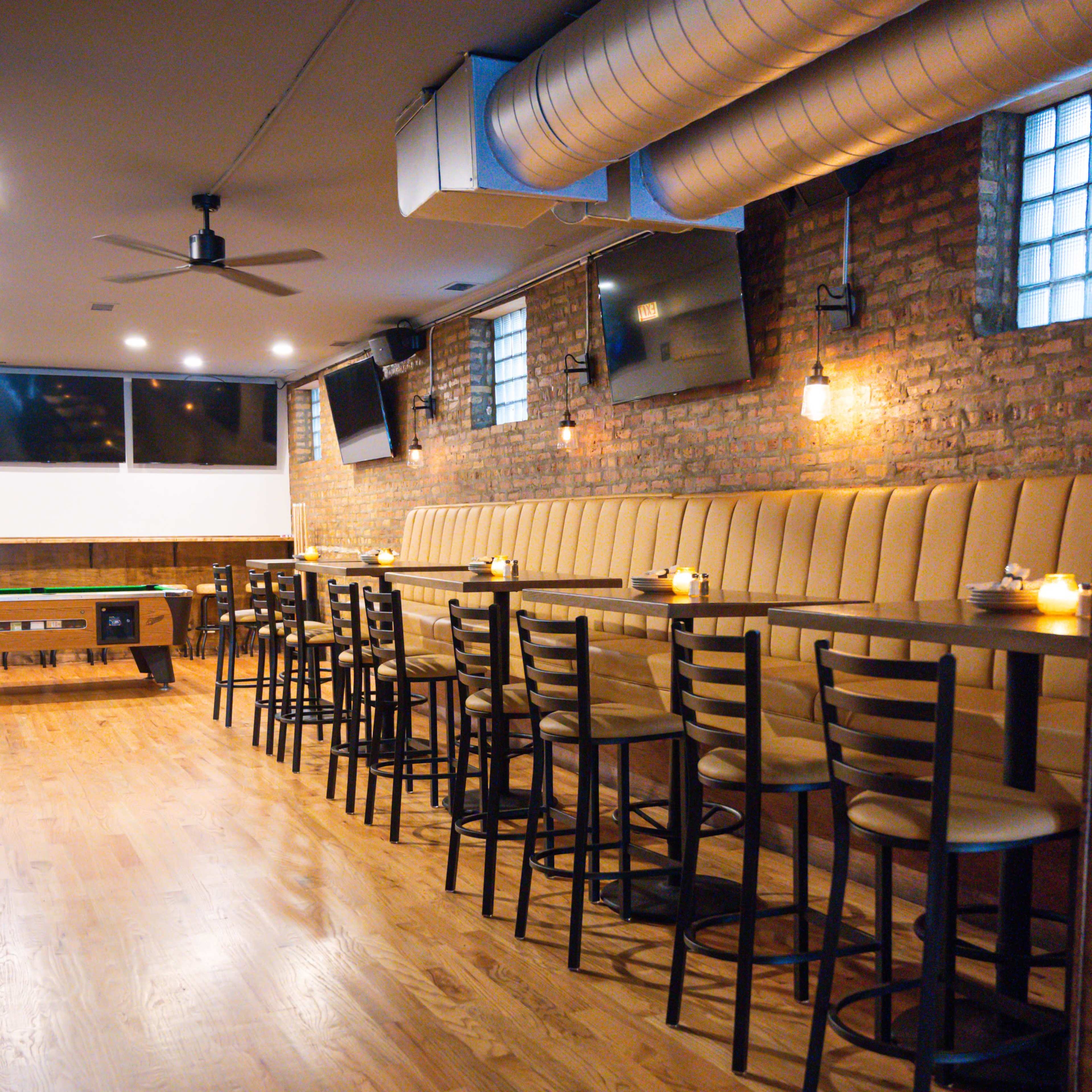 A modern bar interior with a long padded seating area, high tables, and wooden floors, illuminated by pendant lighting.