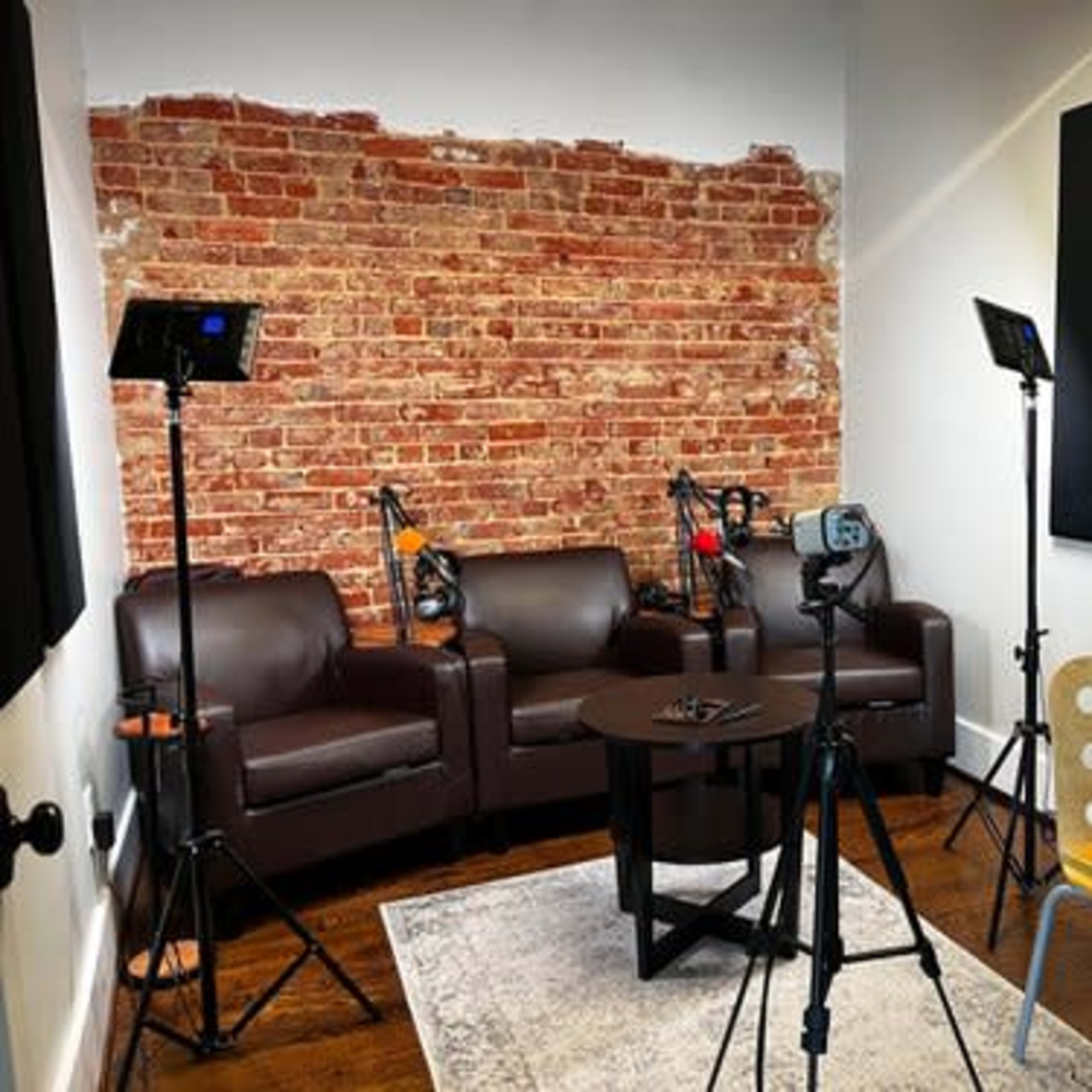A recording studio with brown leather couches, a wooden table, microphones, and lighting equipment against a brick wall.