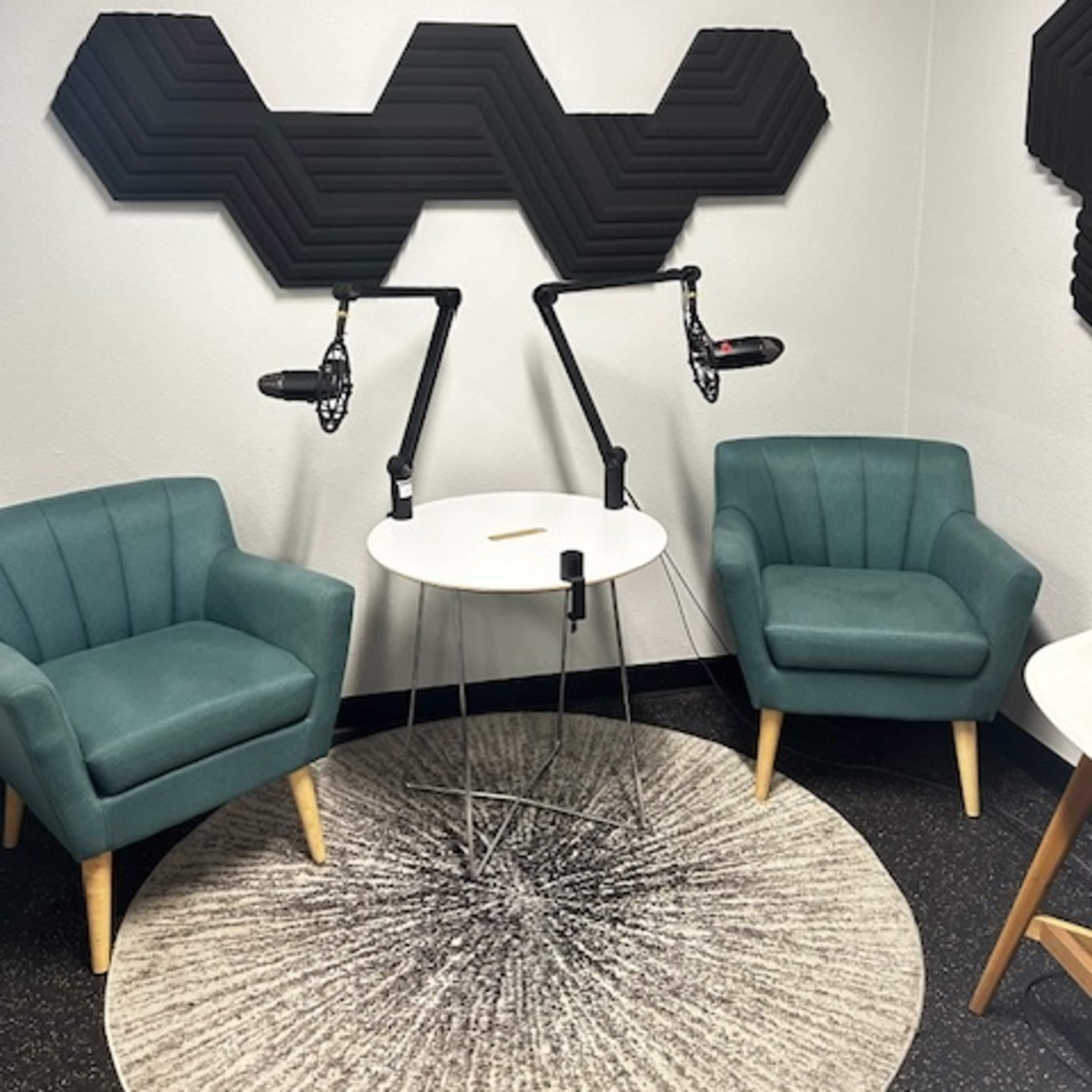 A small recording studio featuring two teal chairs, a round table, and soundproofing panels on the wall above microphone stands.