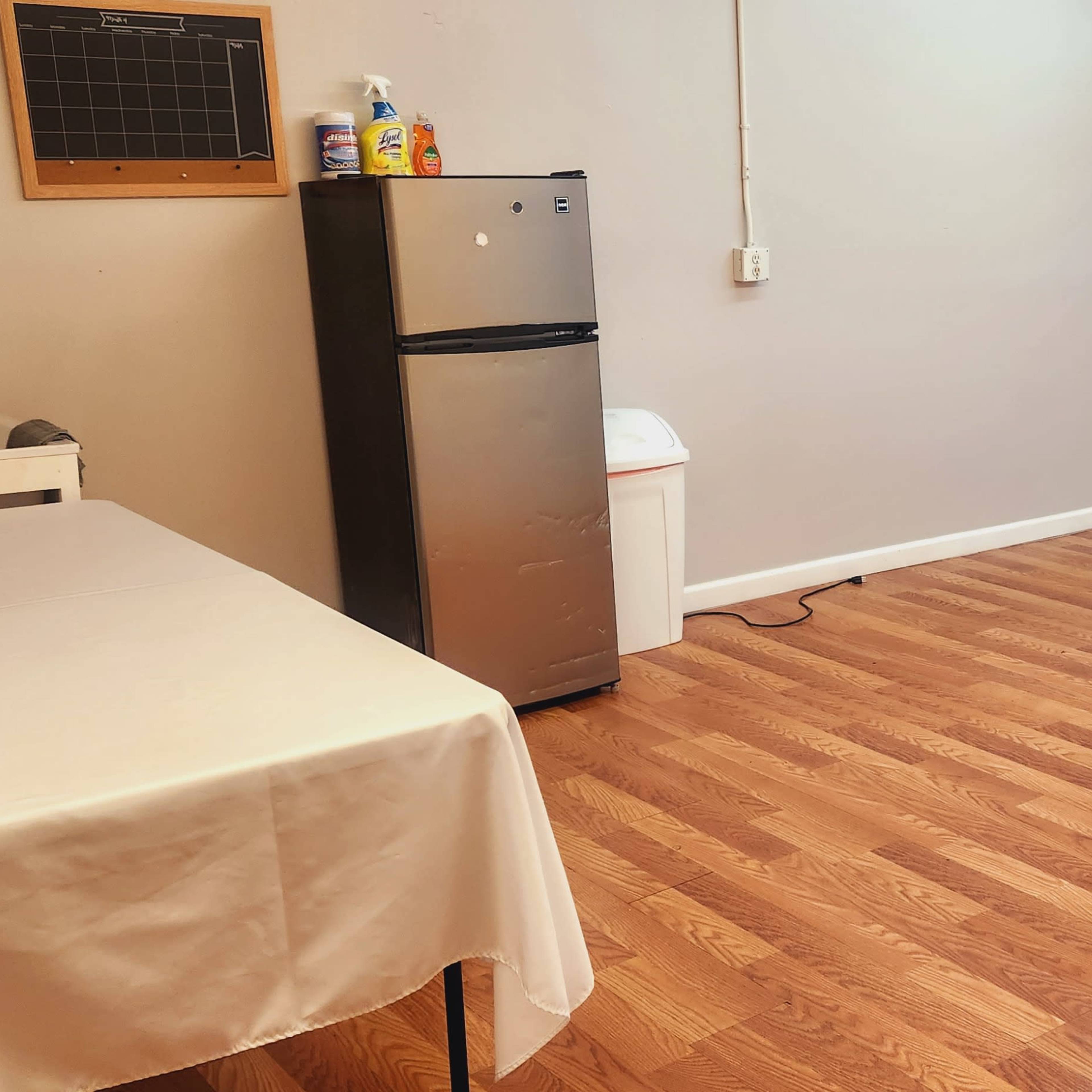 A spacious room with a wooden floor, featuring a table covered with a white cloth, a refrigerator, a trash can, and two chairs against a green wall of foliage.