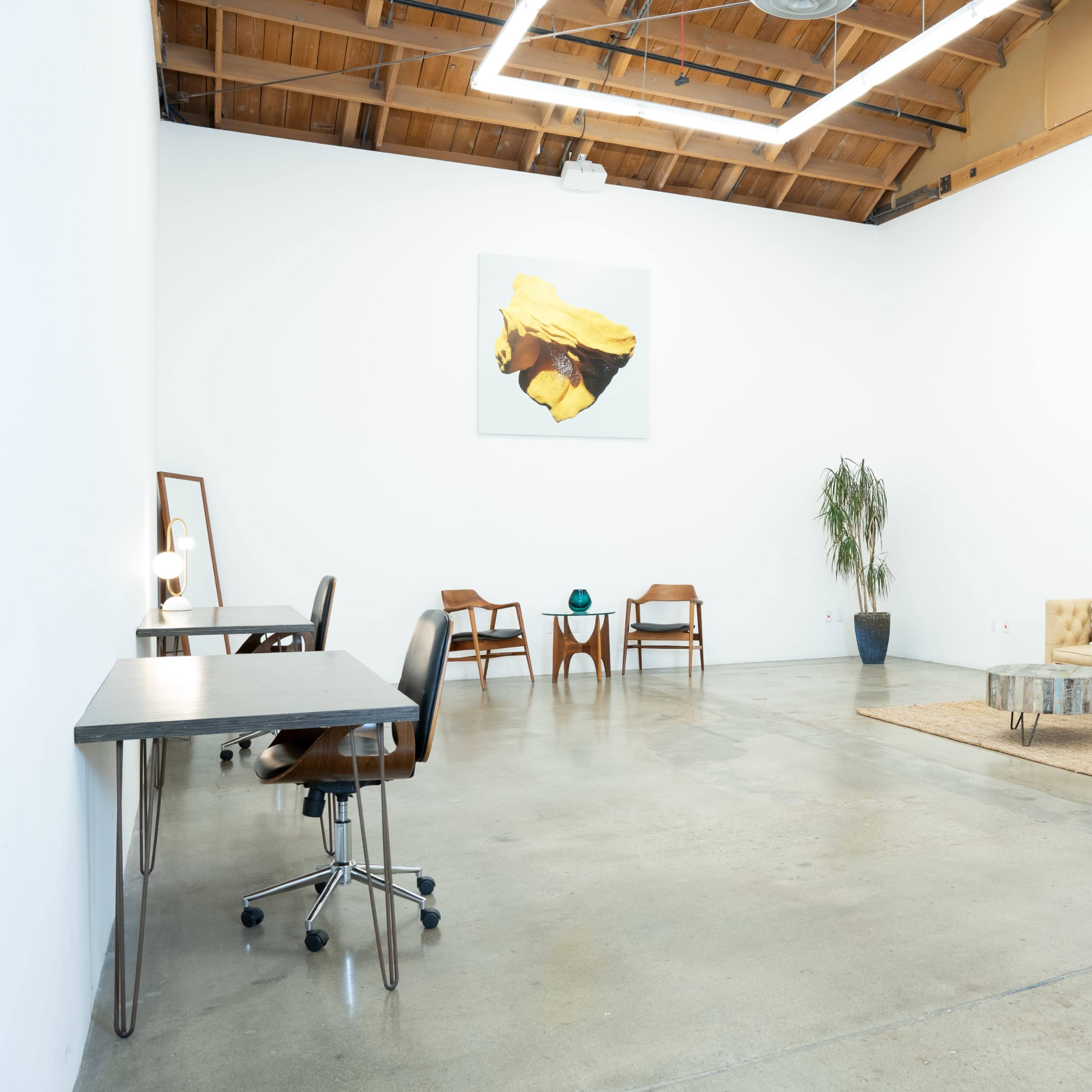 A spacious, minimalist indoor area with a sofa, a coffee table, two desk setups, and a potted plant, all arranged on a concrete floor.
