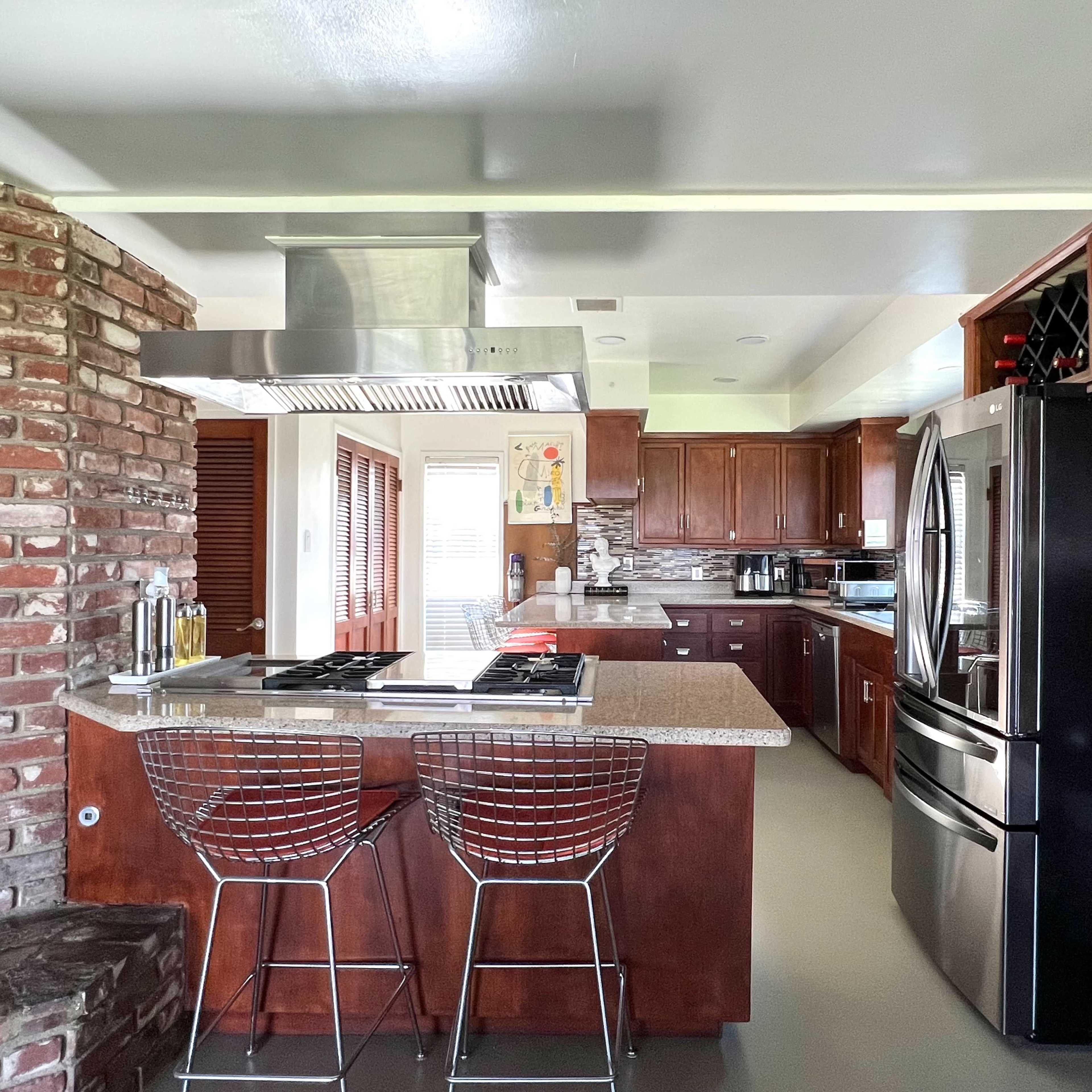 A modern kitchen features wood cabinetry, stainless steel appliances, and a central island with bar stools, adjacent to a brick wall.
