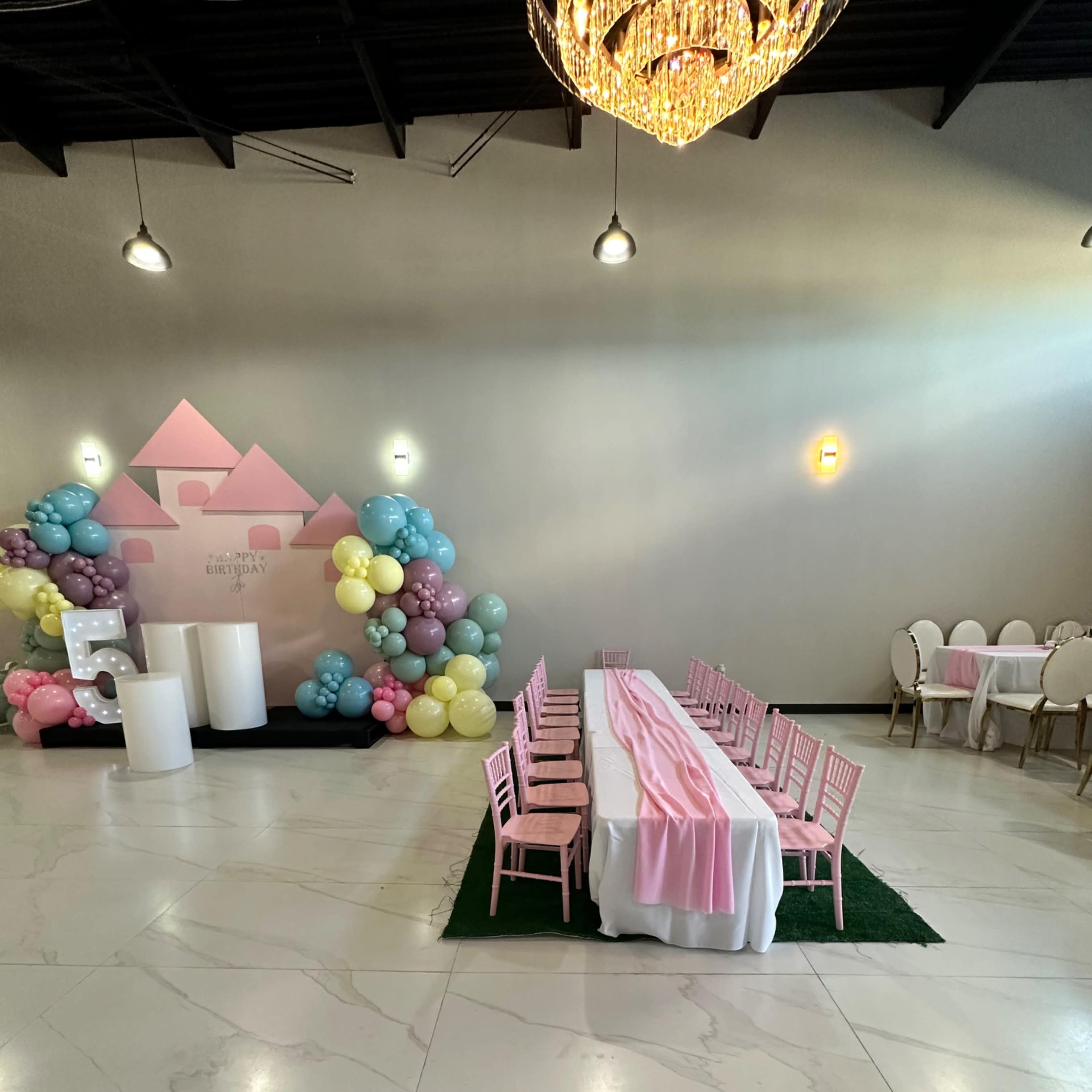 A decorated indoor event space featuring a long table set with pink tablecloths, surrounded by pink chairs, and balloon decorations in pastel colors at one end.