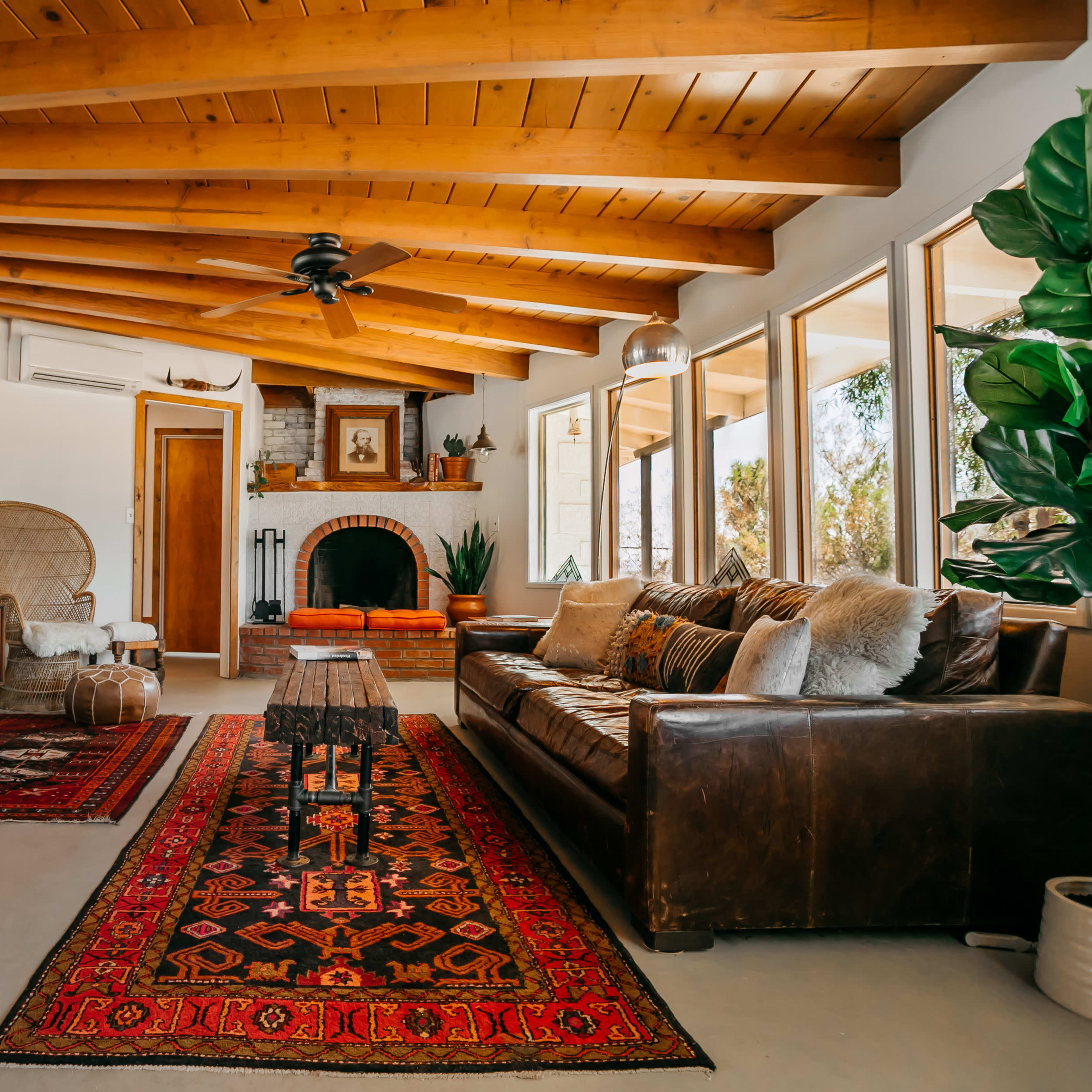A cozy living room features a leather sofa, an area rug, wooden beams on the ceiling, and large windows with views of greenery.