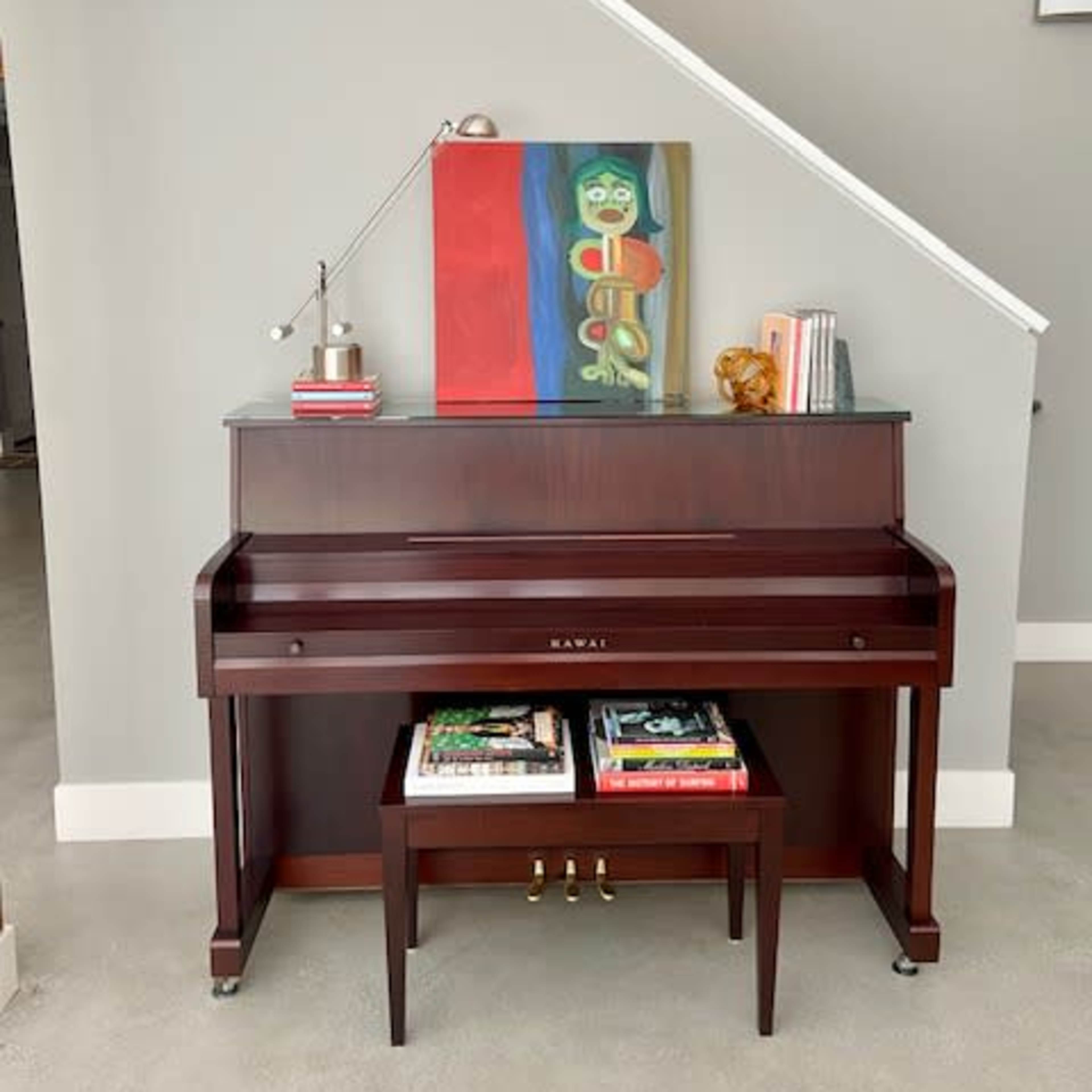 A brown piano is positioned against a light gray wall, with a colorful painting and decorative items placed on top, and magazines stored underneath.