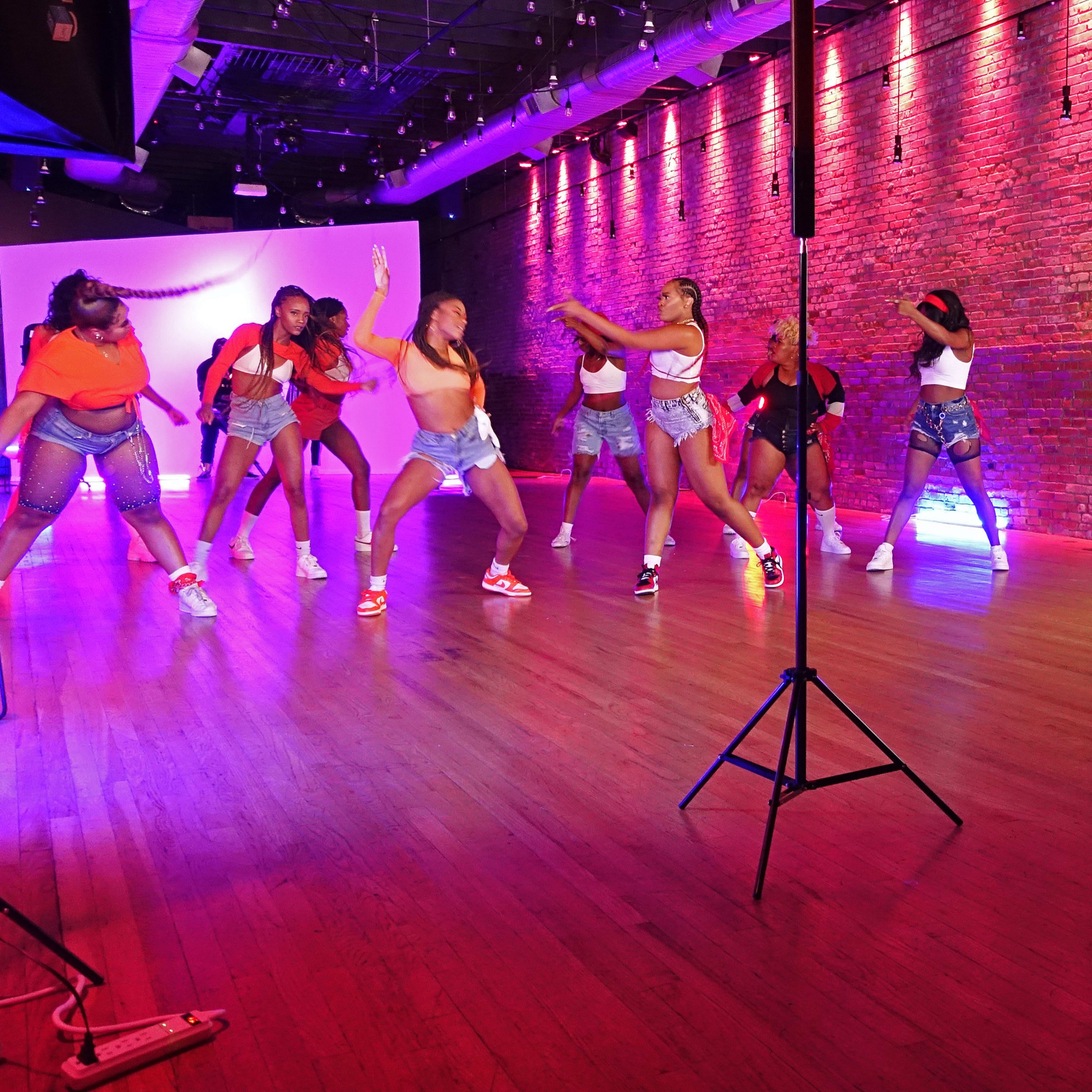 A group of seven dancers performs a choreographed routine in a studio with vibrant lighting and a photographer capturing the scene.