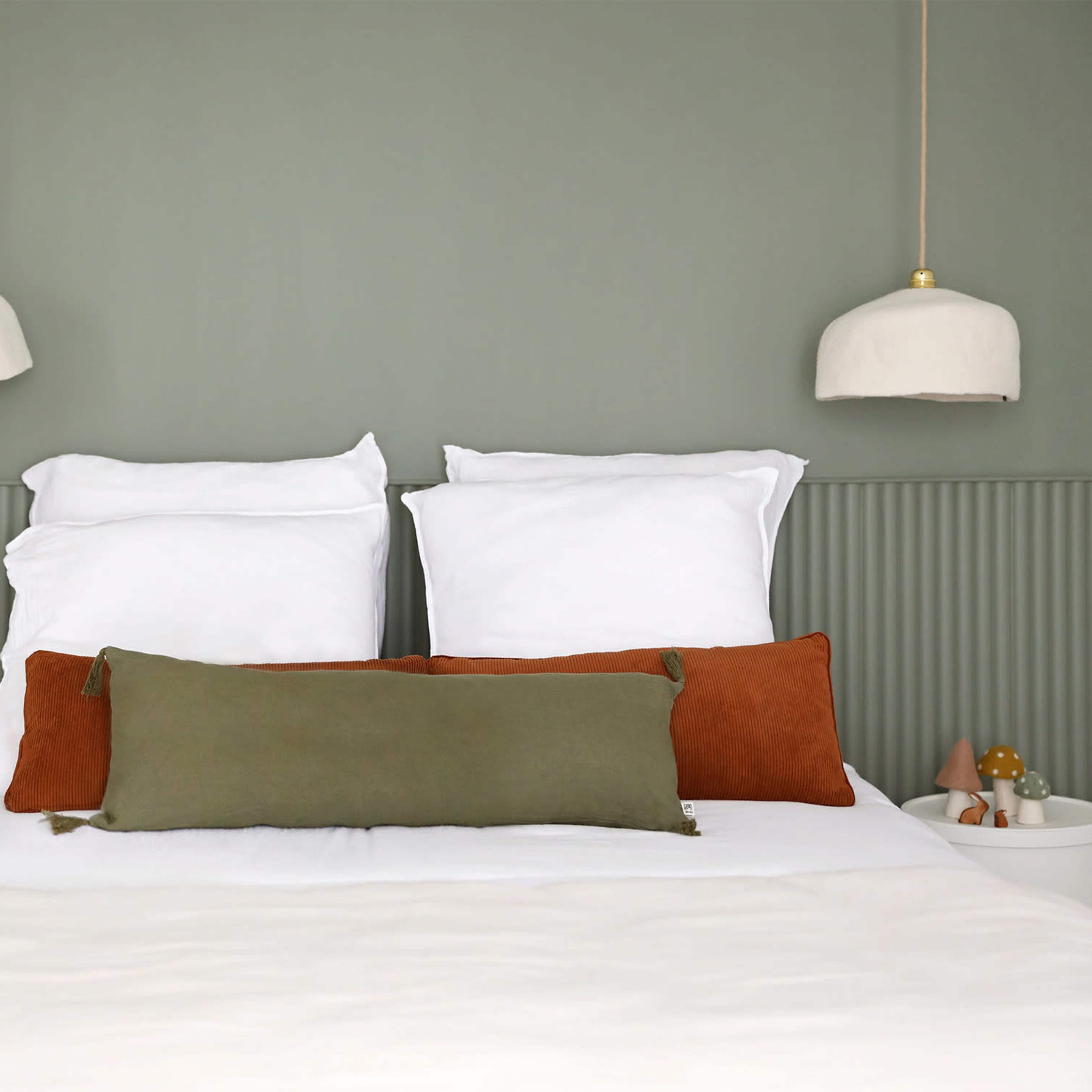 A neatly made bed with white linens, two white pillows, and decorative cushions is positioned against a green wall, flanked by pendant lights.