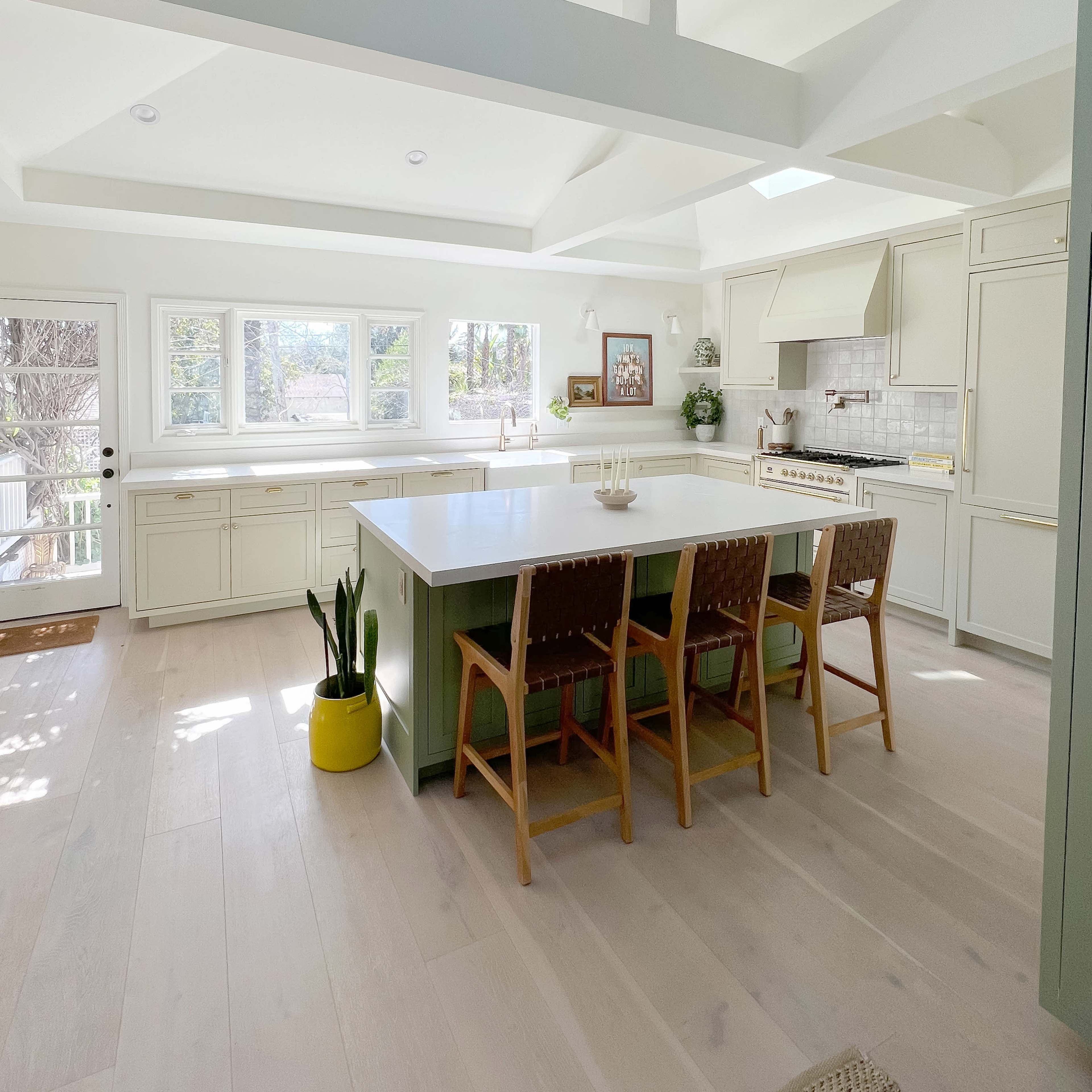 A modern kitchen features a large island with wooden stools, bright lighting, and windows overlooking a natural landscape.