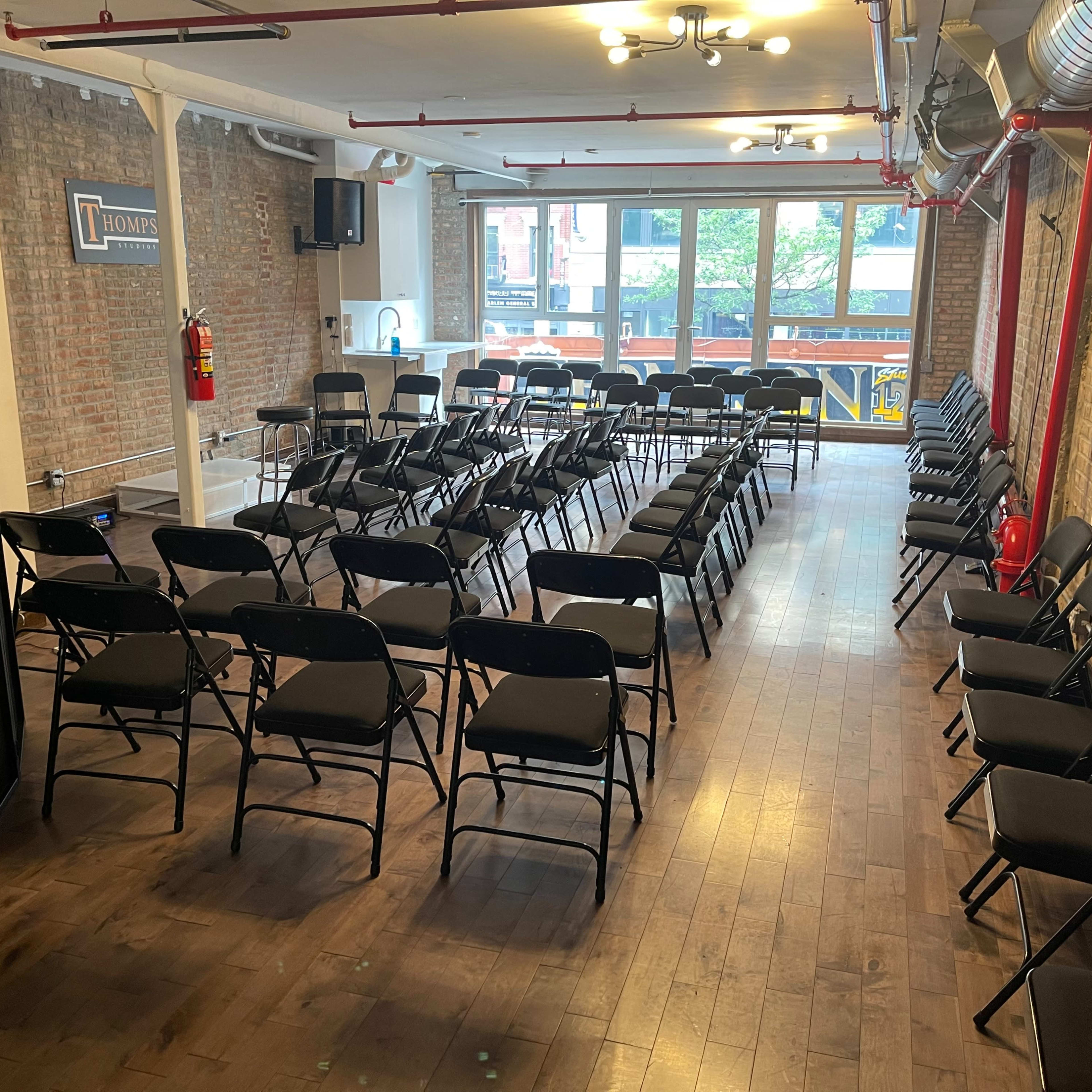 The room features multiple rows of black folding chairs arranged facing a large window, with exposed brick walls and wooden flooring.