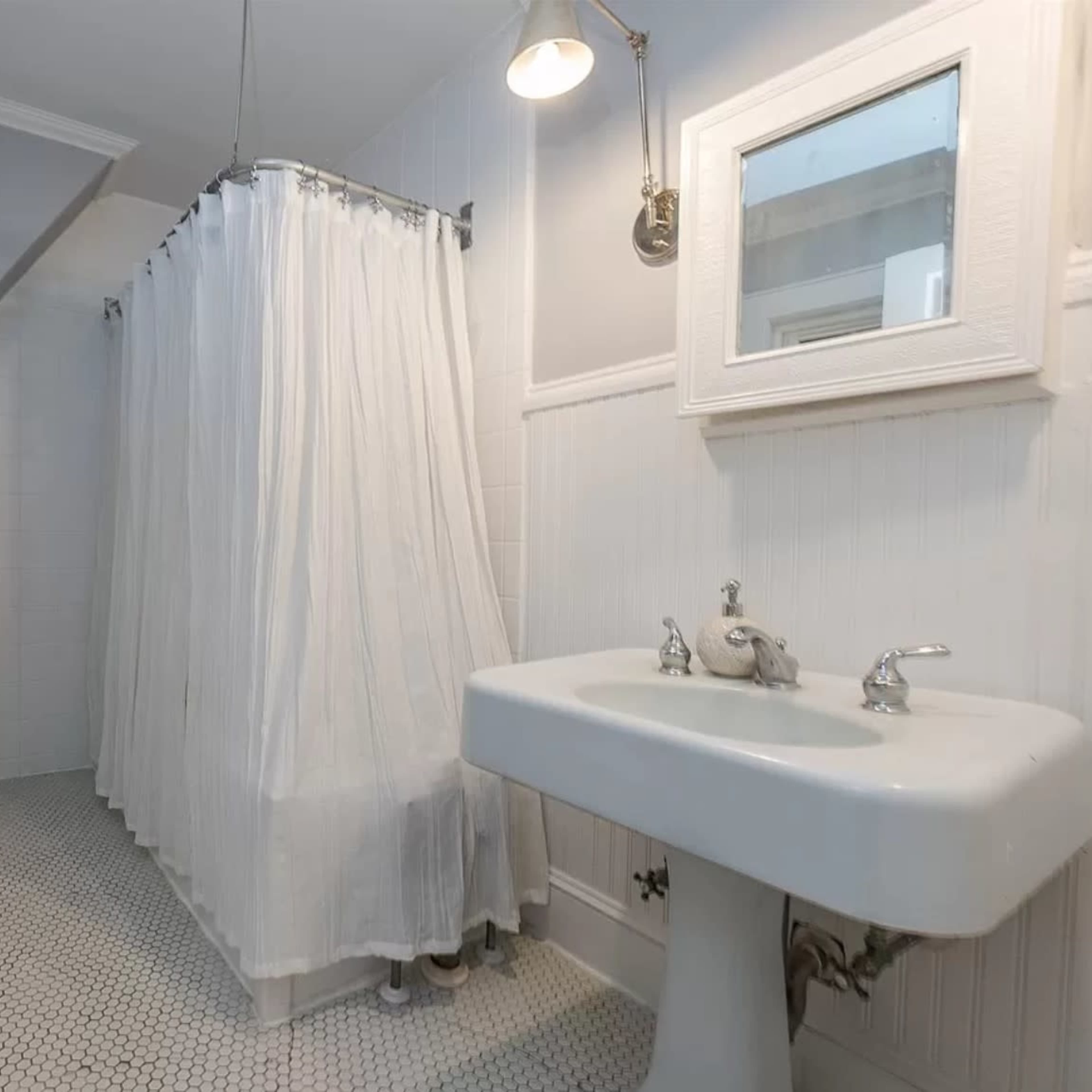 A bathroom with a toilet, a shower area with a curtain, and a sink, all set against light-colored walls and hexagonal tile flooring.