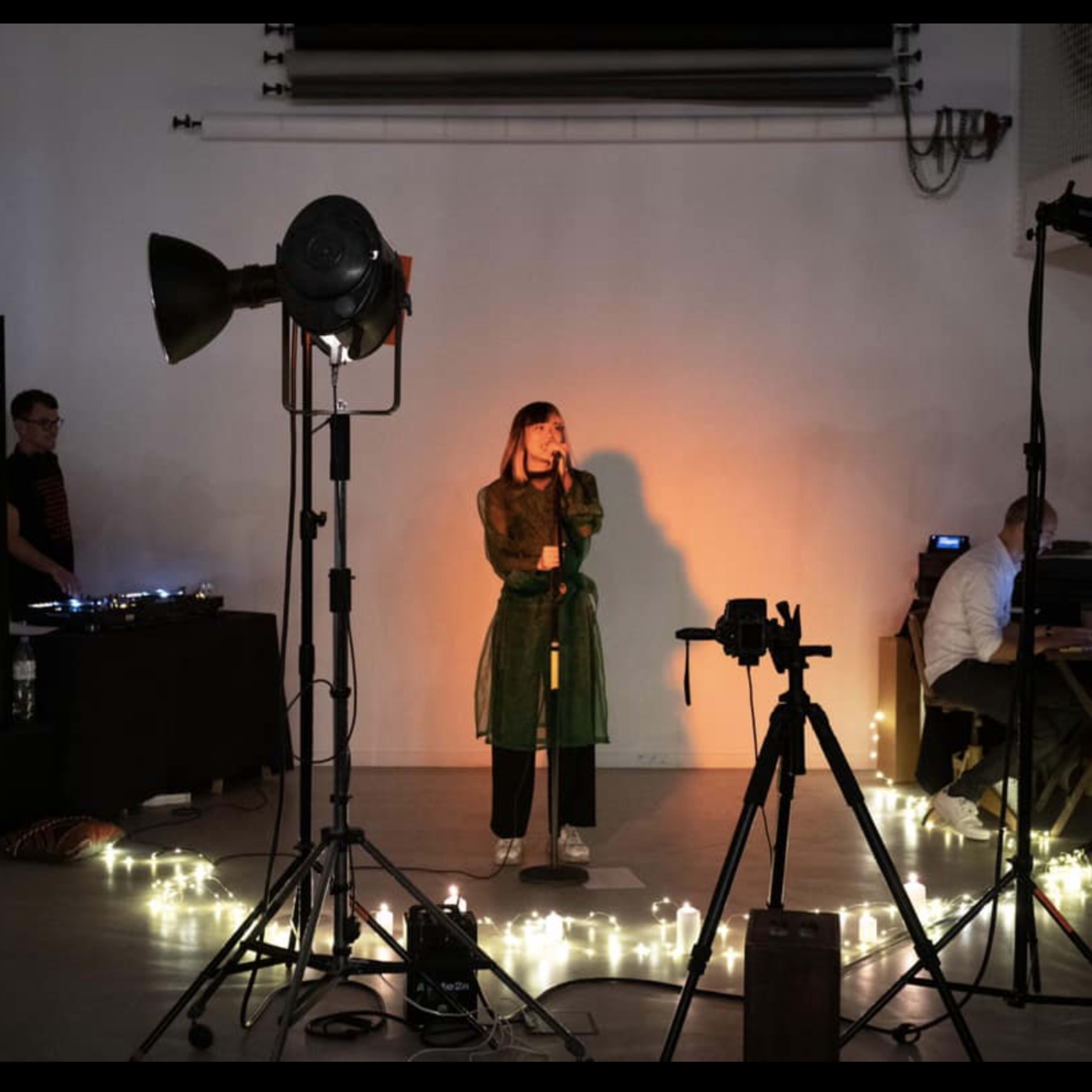 A woman stands on a stage with a microphone, surrounded by lights and camera equipment while a DJ performs in the background.