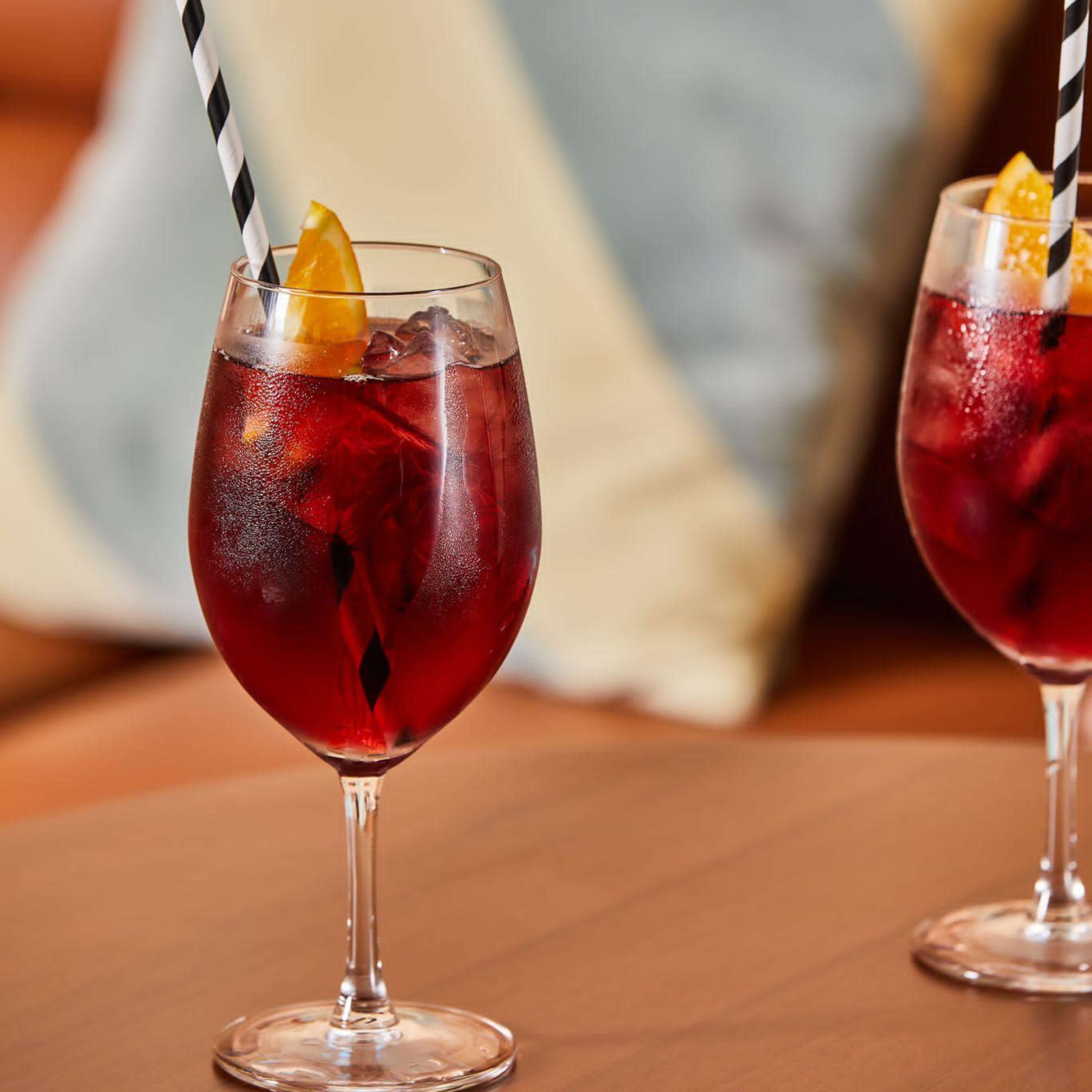 Two glasses of a red beverage with ice and a slice of orange sit on a wooden table, with a striped straw in each glass, beside a patterned cushion in the background.