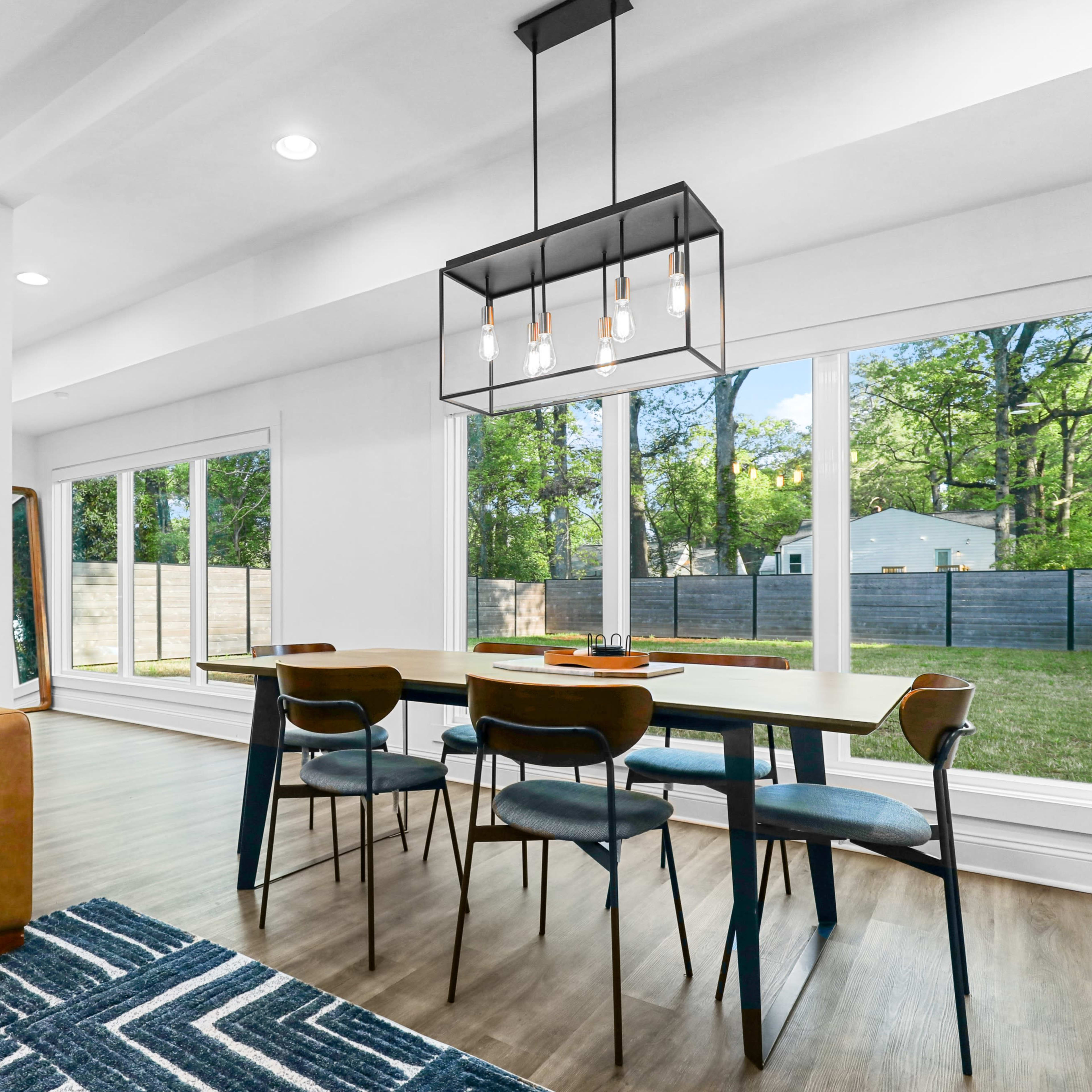 A modern dining area features a large table surrounded by chairs, with a brown sofa and large windows overlooking a green backyard.
