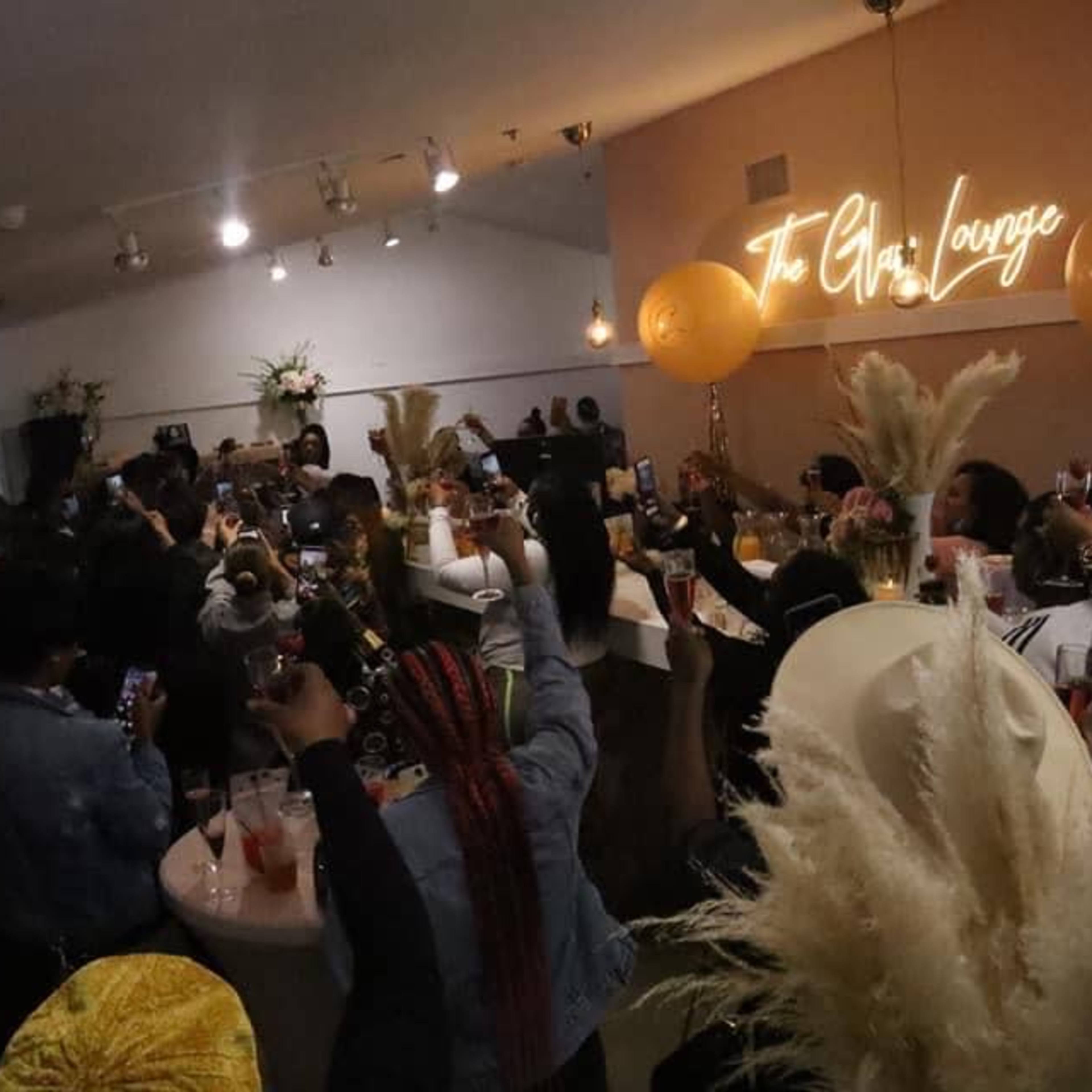 A crowd of people raises their drinks in celebration inside a lounge with a glowing sign that reads "The GvL Lounge."