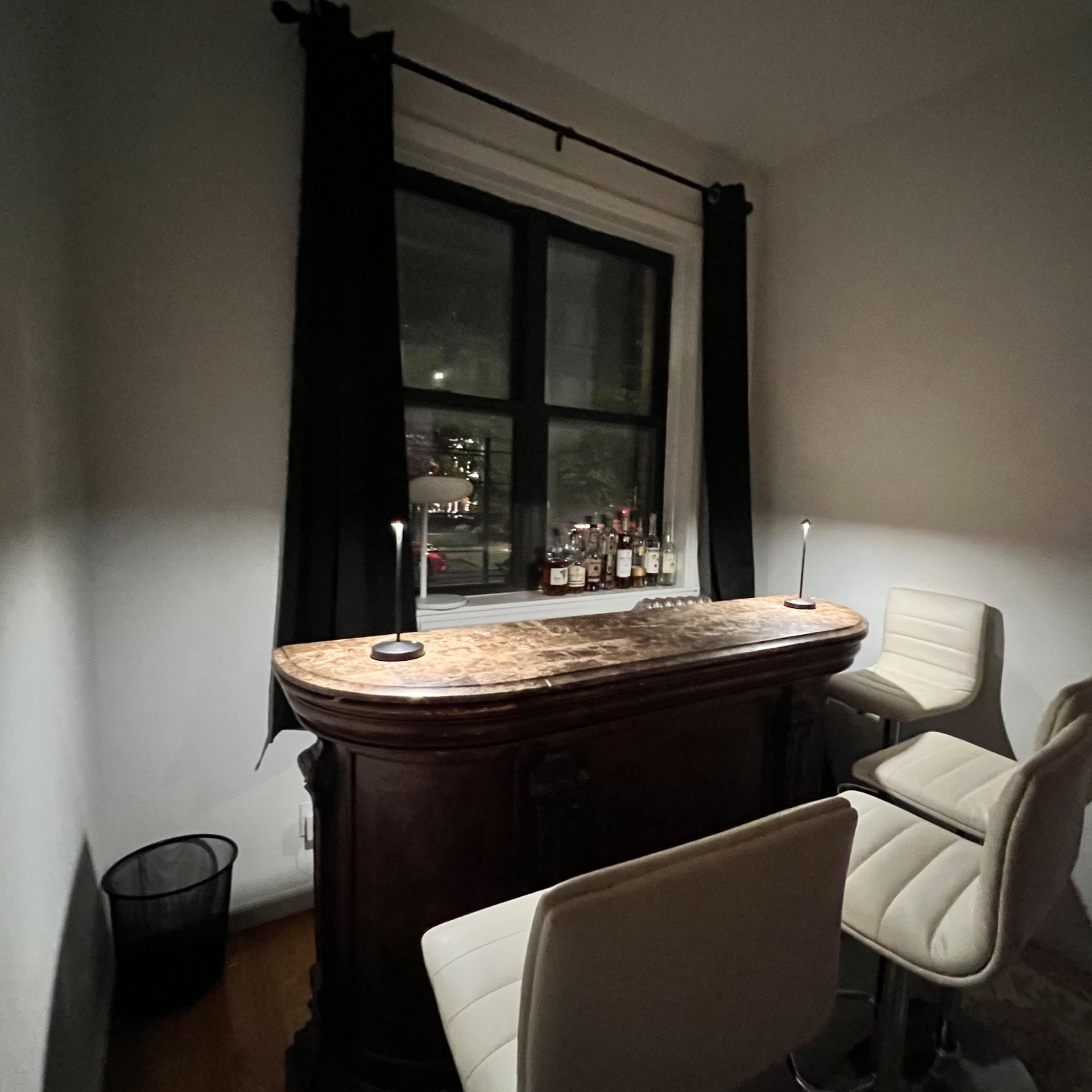 A small home bar area features a wooden bar top with two white stools, illuminated by two lamps, and a window visible in the background.