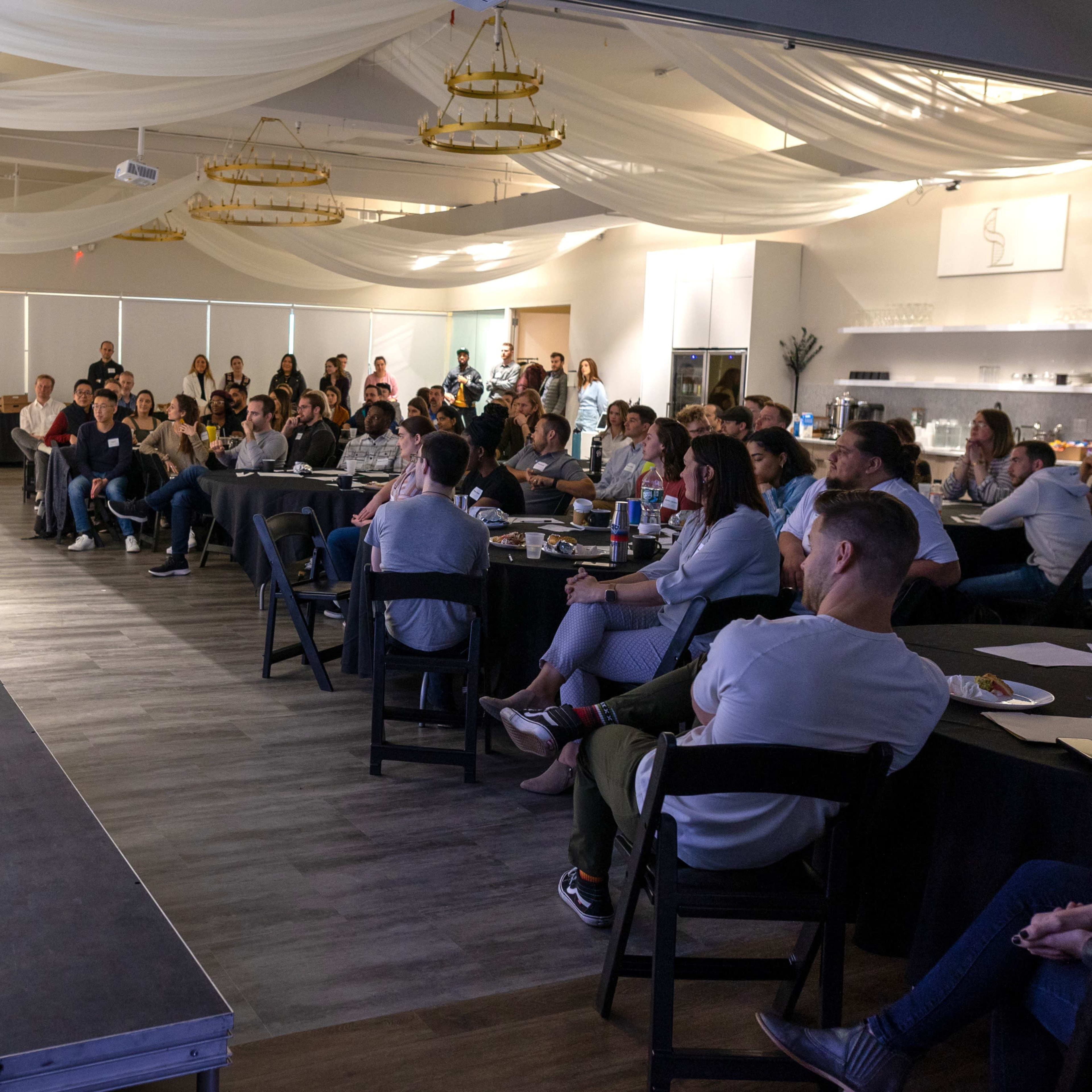 A speaker presents to an audience seated at tables in a well-lit event space with decorative elements.