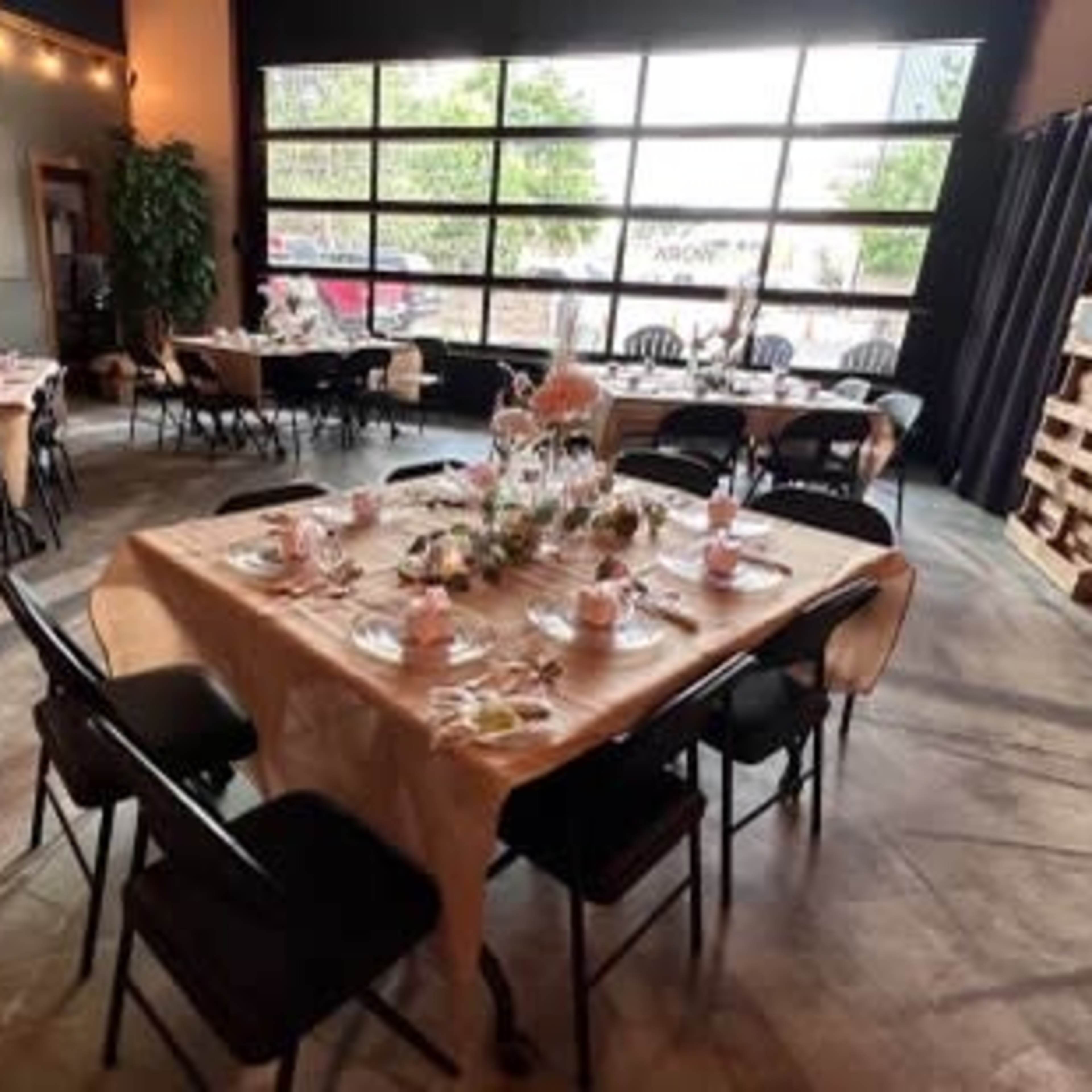 A dining area arranged with several tables covered in beige tablecloths, set for a meal with chairs around them and large windows in the background.